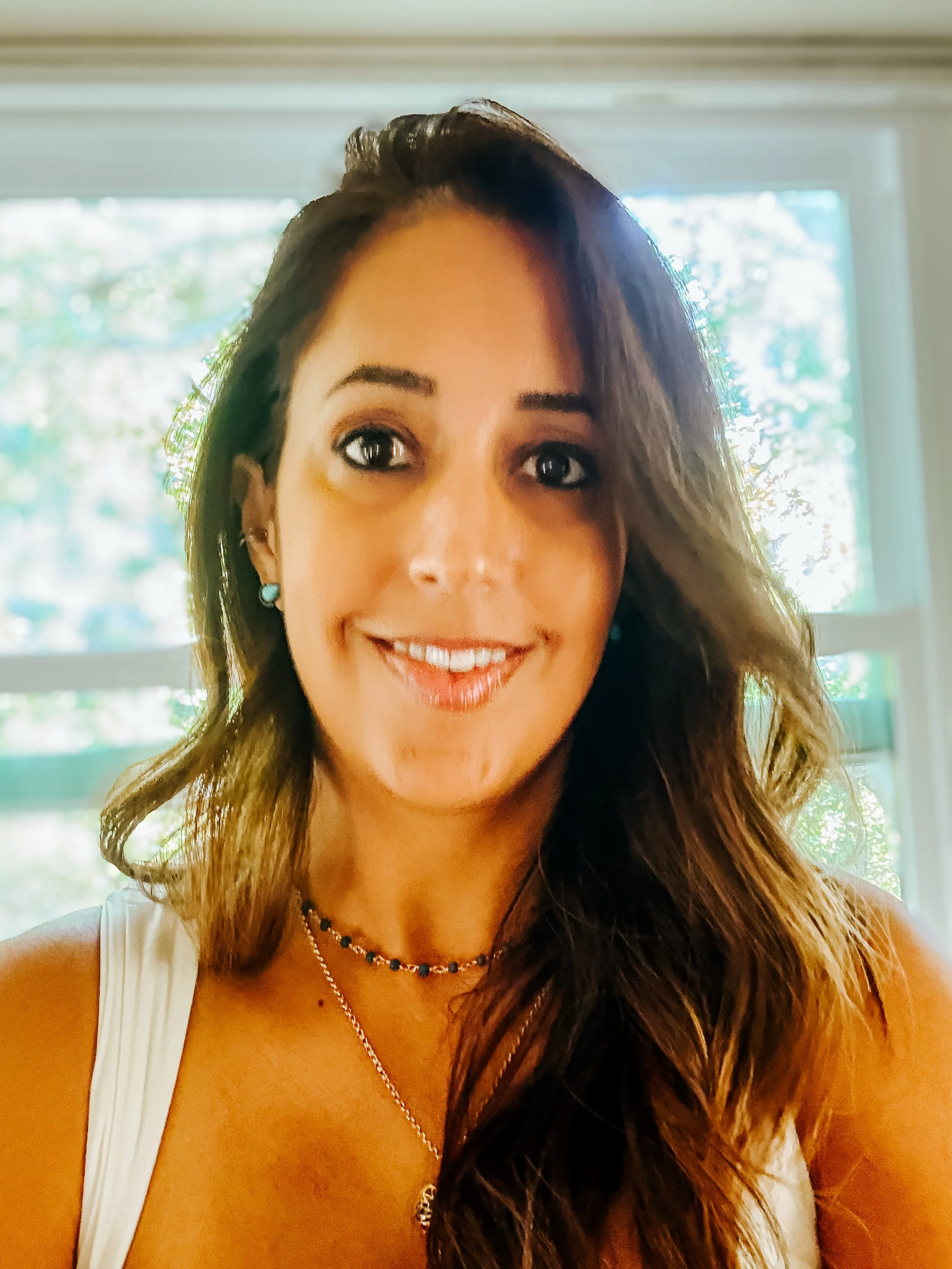 A woman with wavy brown hair, wearing earrings and jewelry, smiling indoors with a window in the background.