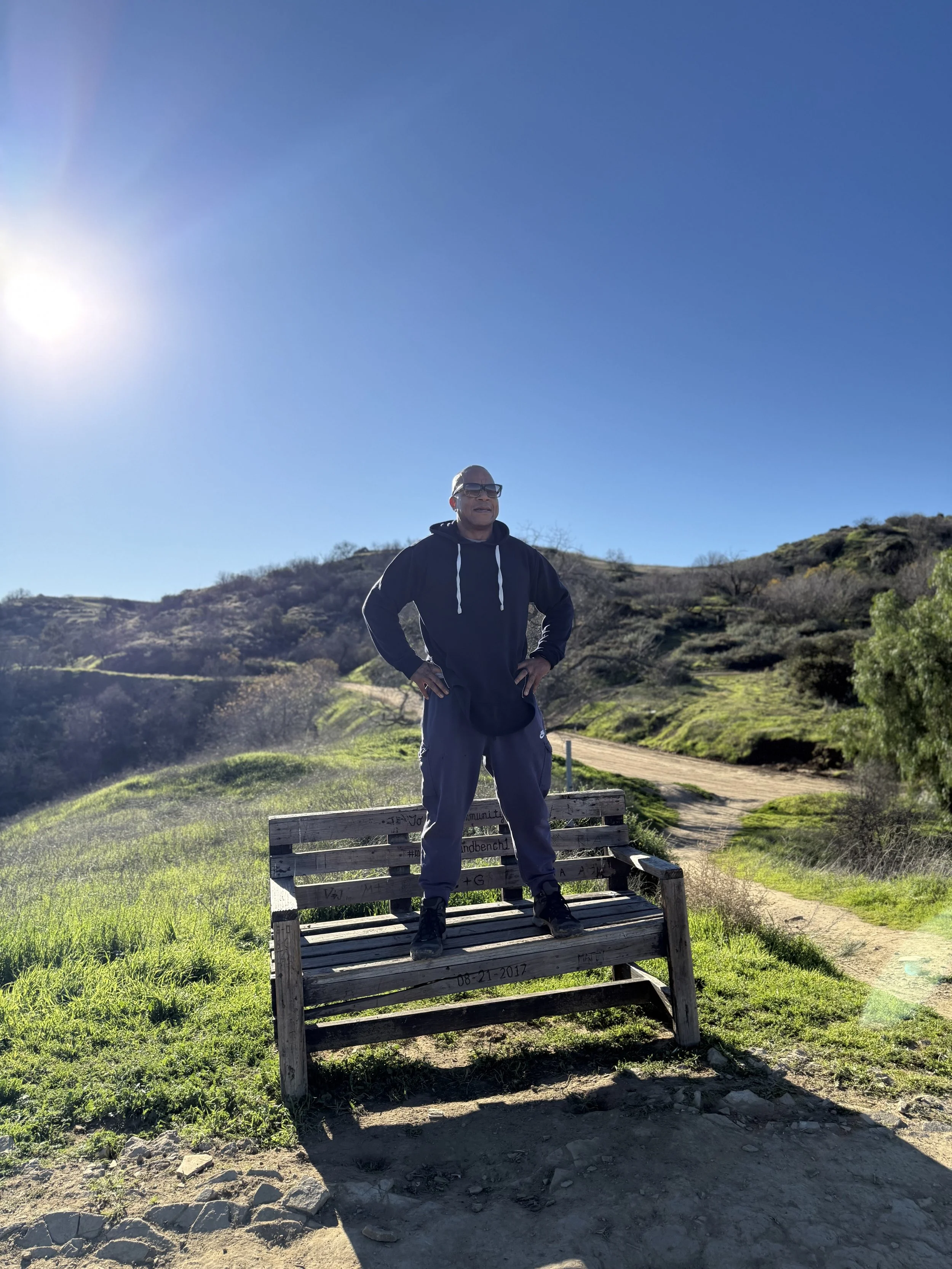 A man stands on a wooden bench in a scenic outdoor setting with green hills, a dirt path, and a bright blue sky with the sun shining.