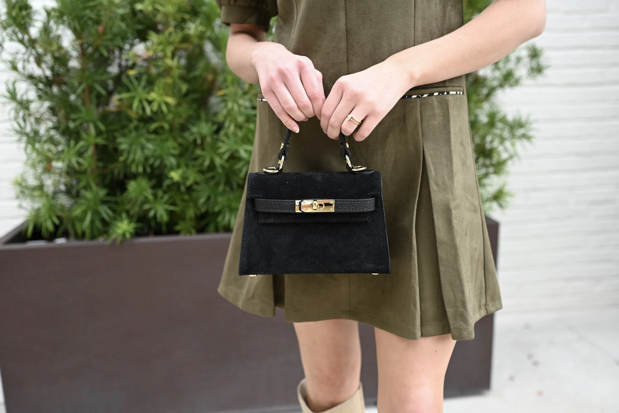 A woman holding a black handbag with a gold clasp, standing outdoors in front of green plants and a white wall.