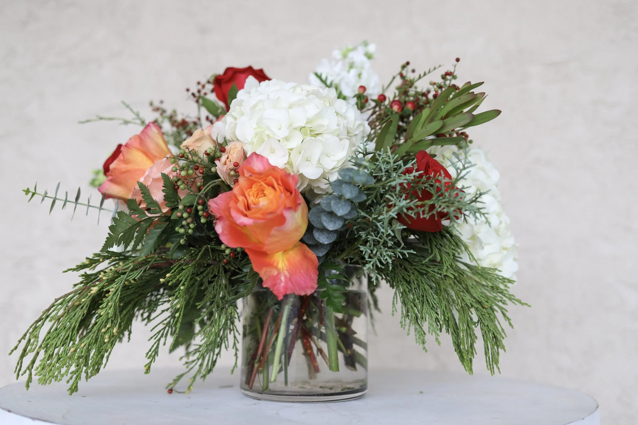 A floral arrangement in a glass vase contains white, orange, and red roses, green foliage, and sprigs of other greenery.