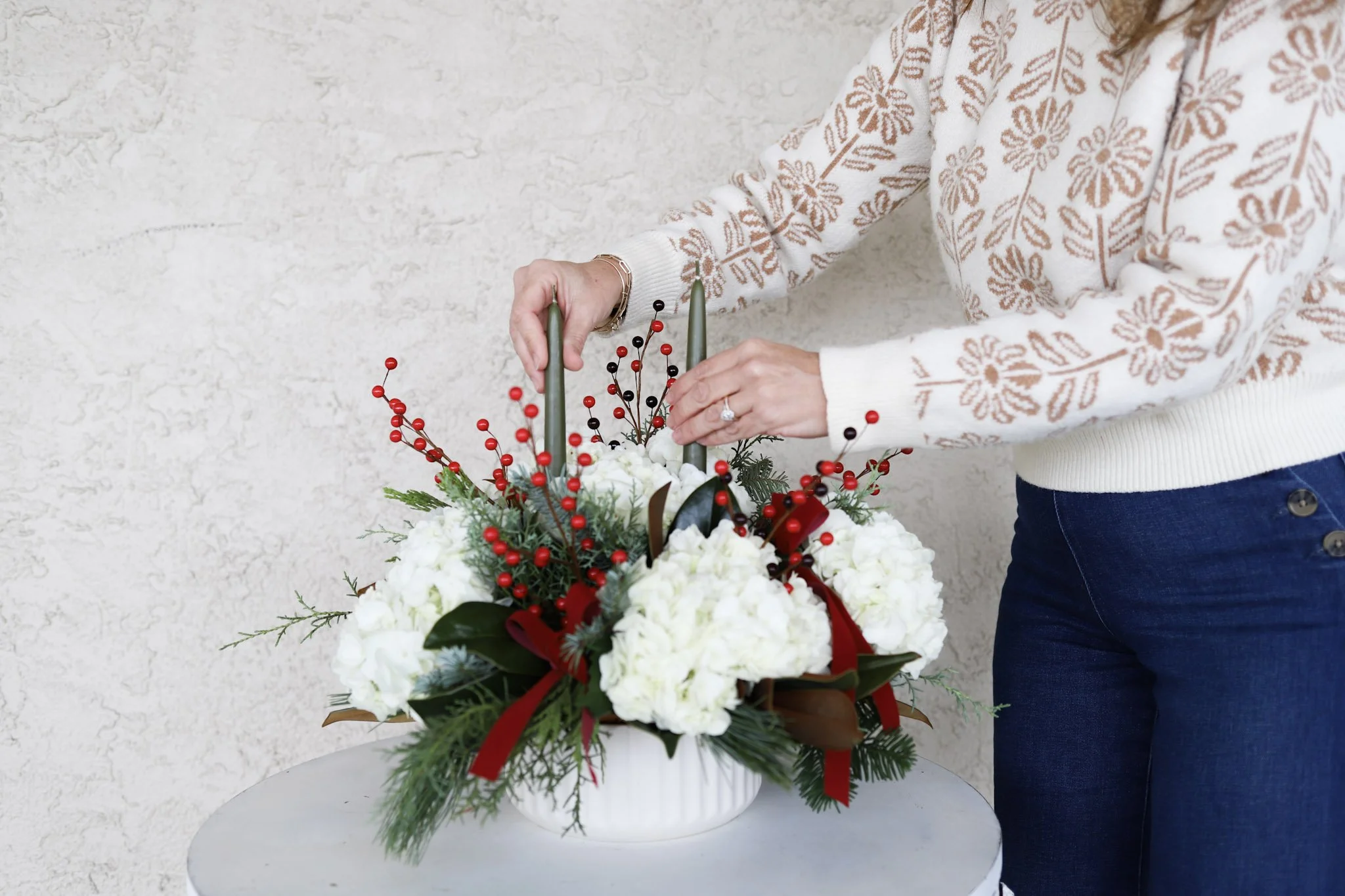 Person arranging a holiday floral centerpiece with white flowers, red berries, and greenery on a white round table against a textured light-colored wall.