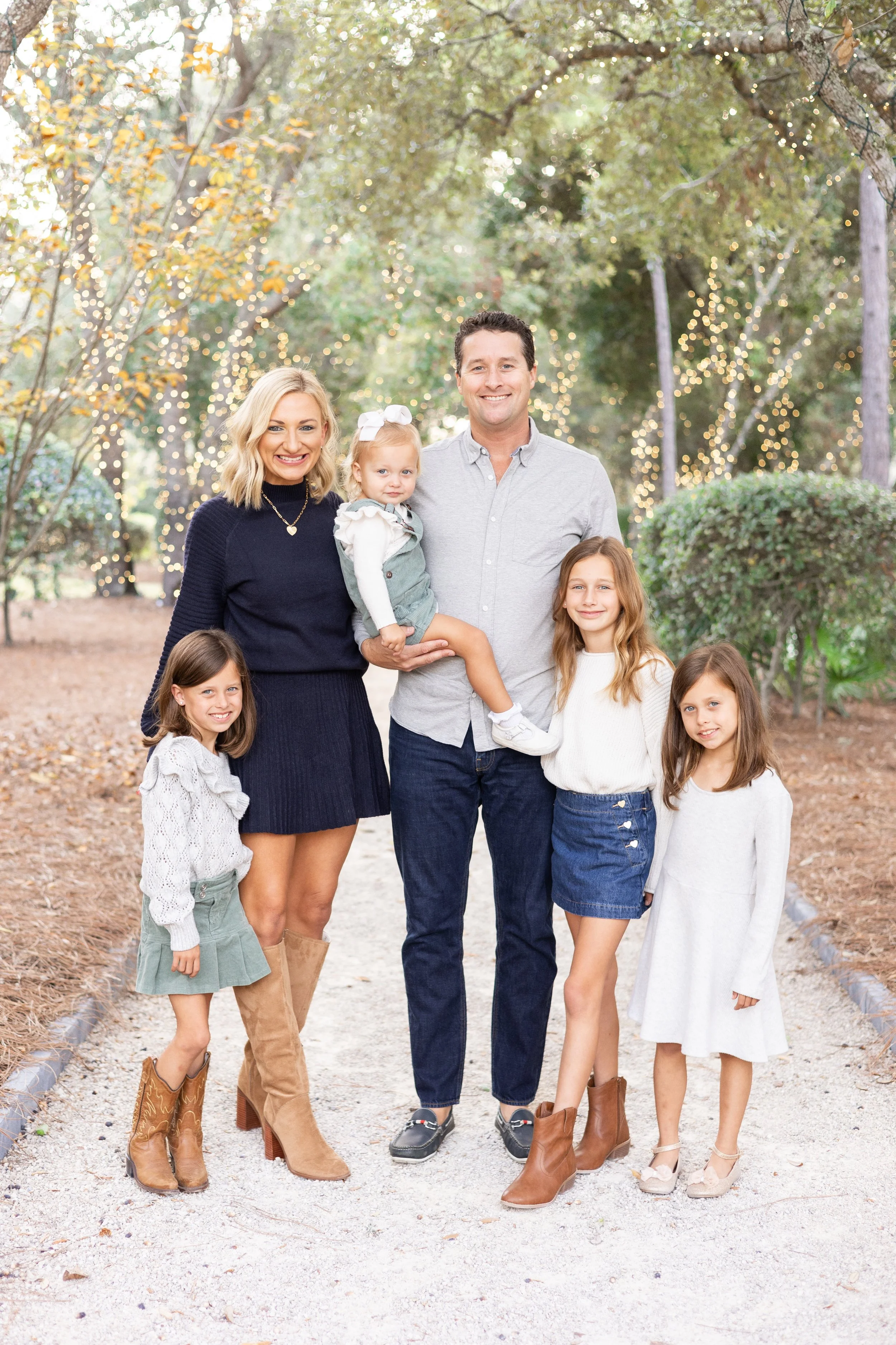 Family of six, three adult women and three young girls, standing outdoors on a trail with trees and twinkle lights in the background. The adults are smiling and the girls are dressed in casual fall attire.