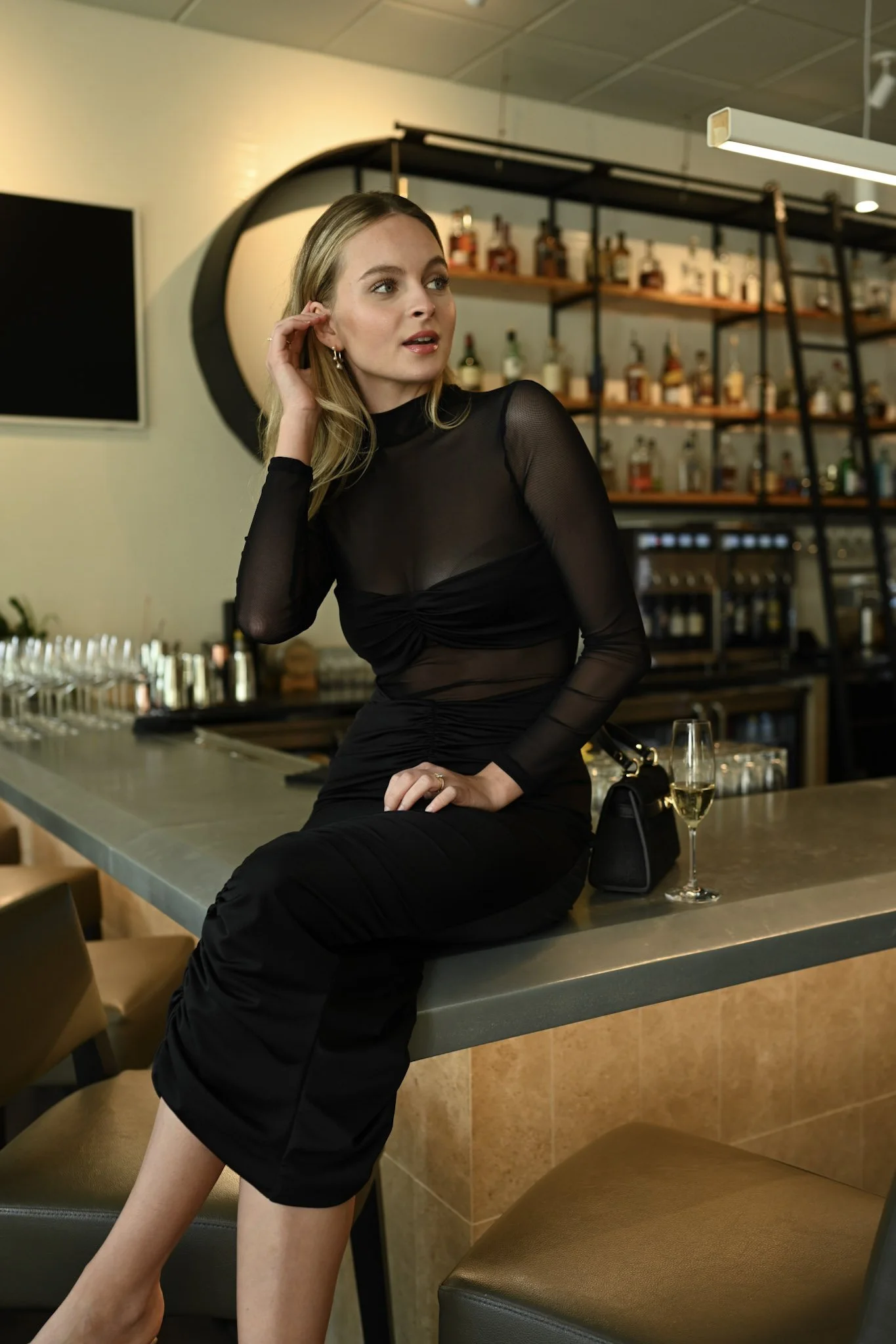 A woman in a black, sheer, long sleeve dress with a high neck, sitting on a bar counter with a glass of champagne beside her, in a bar or lounge setting with shelves of bottles in the background.