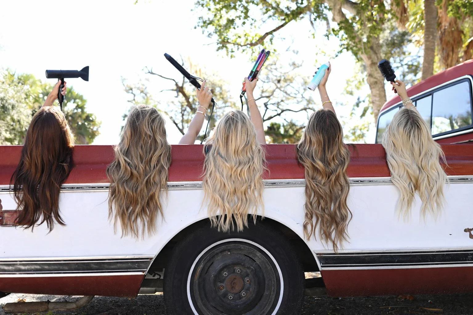 Five women are sitting in the bed of a vintage red and white pickup truck, with long, wavy hair hanging over the side. They are holding hair styling tools and sprays, suggesting they are doing hair or enjoying a fun outing in a natural outdoor setting.
