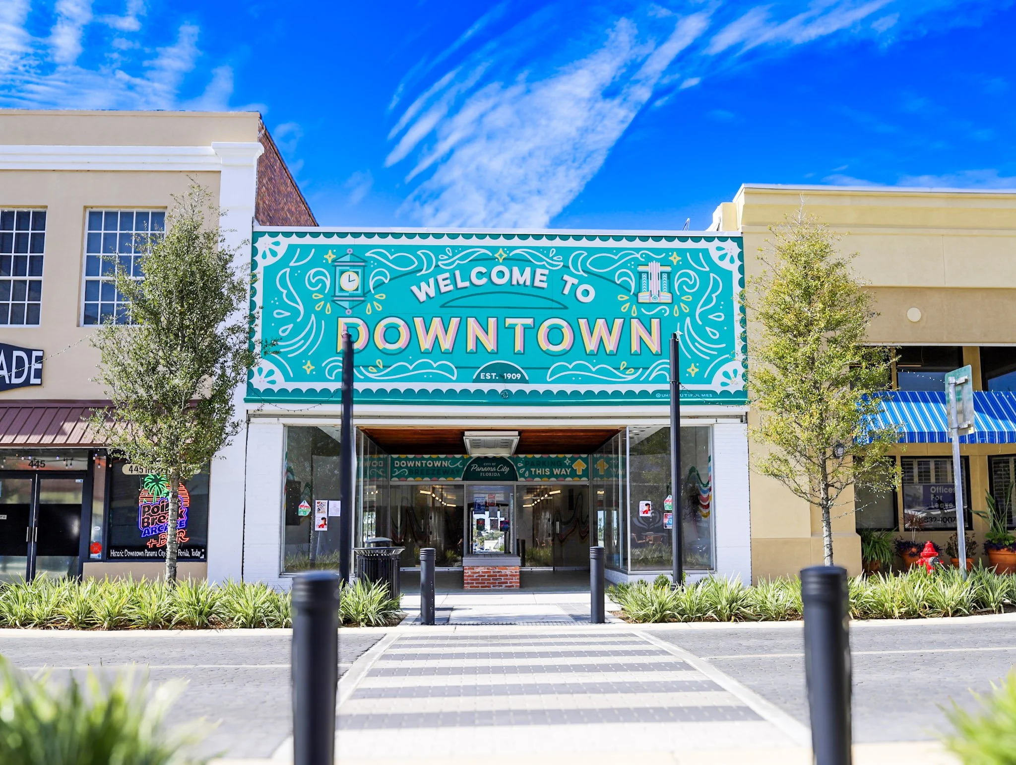 Colorful sign reading 'Welcome to Downtown' above a shopping mall entrance, flanked by trees and a clear blue sky.