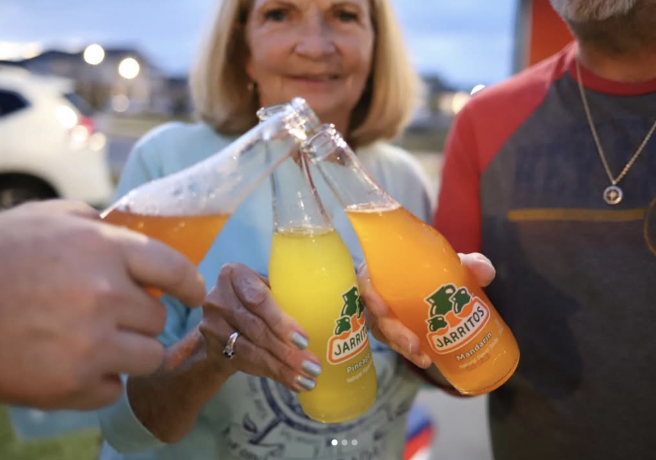 People clinking glass bottles of Jarritos soda, including pineapple and mandarin flavors, outdoors in the evening.