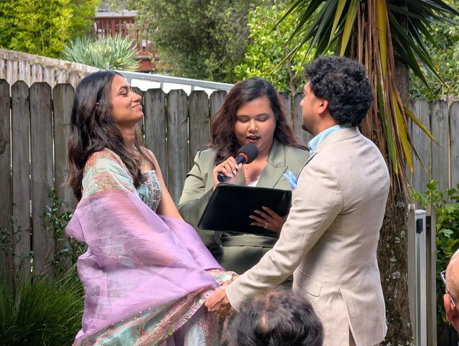 A couple is holding hands during a wedding ceremony outdoors, with a Alison Jimmy, the officiant reading from a tablet and holding a microphone.