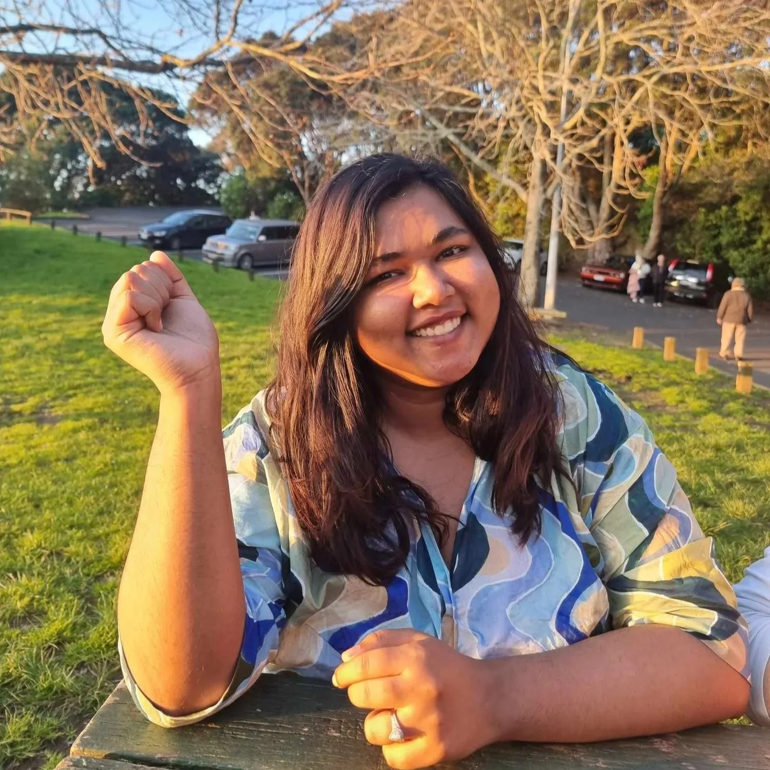 Alison Jimmy with shoulder-length dark hair smiling in an outdoor park during sunset, with trees and parked cars in the background.