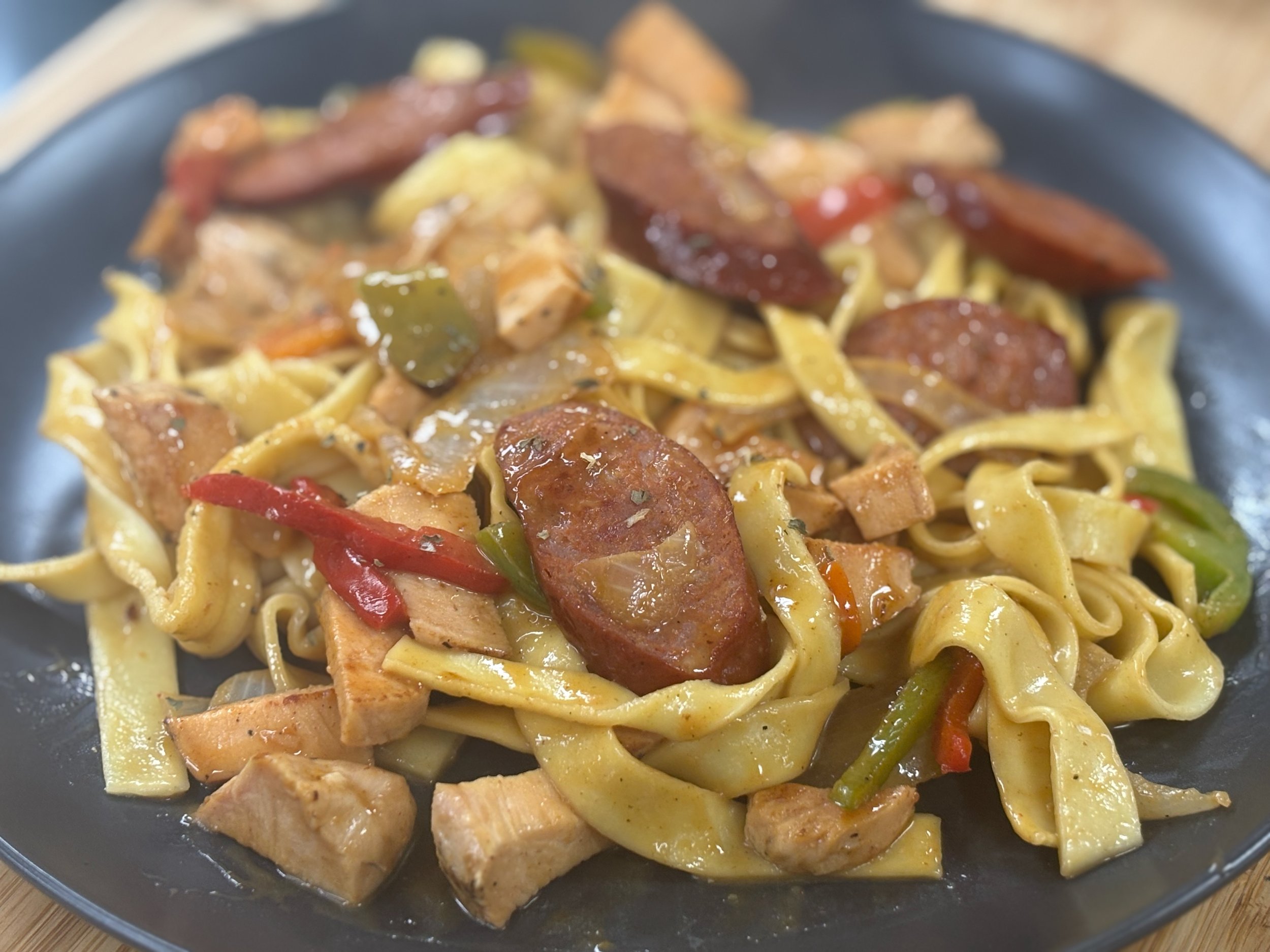 Plate of pasta with sliced sausage, chicken, and vegetables in a savory sauce.