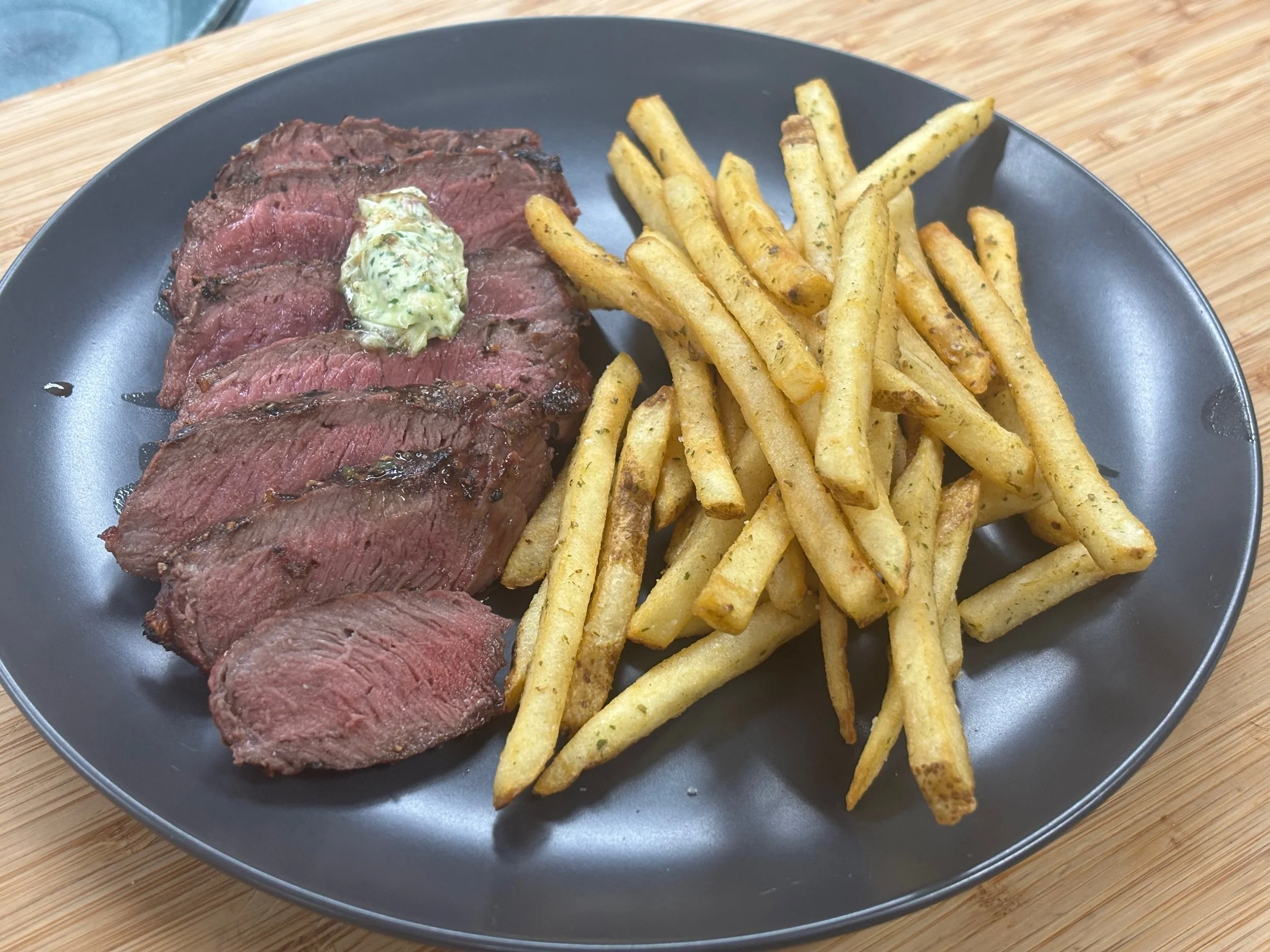 Plate of medium-rare steak with butter on top and a side of seasoned French fries on a black plate.