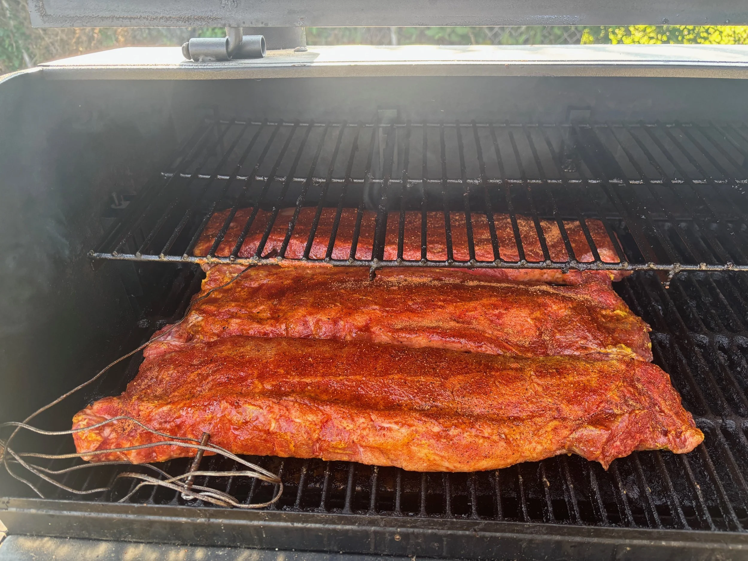 Two racks of seasoned pork ribs cooking on a black outdoor grill, with a second grill rack above.