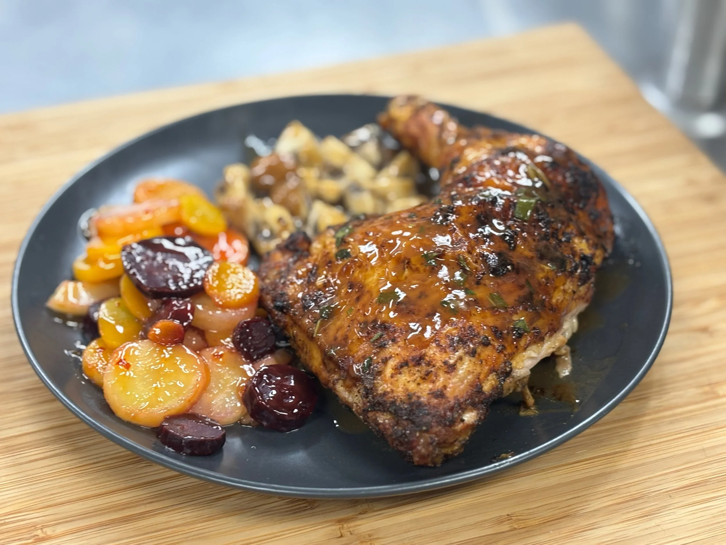 A plate of roasted chicken leg with glazed carrots, cherries, and a side of mashed potatoes with gravy on a wooden surface.