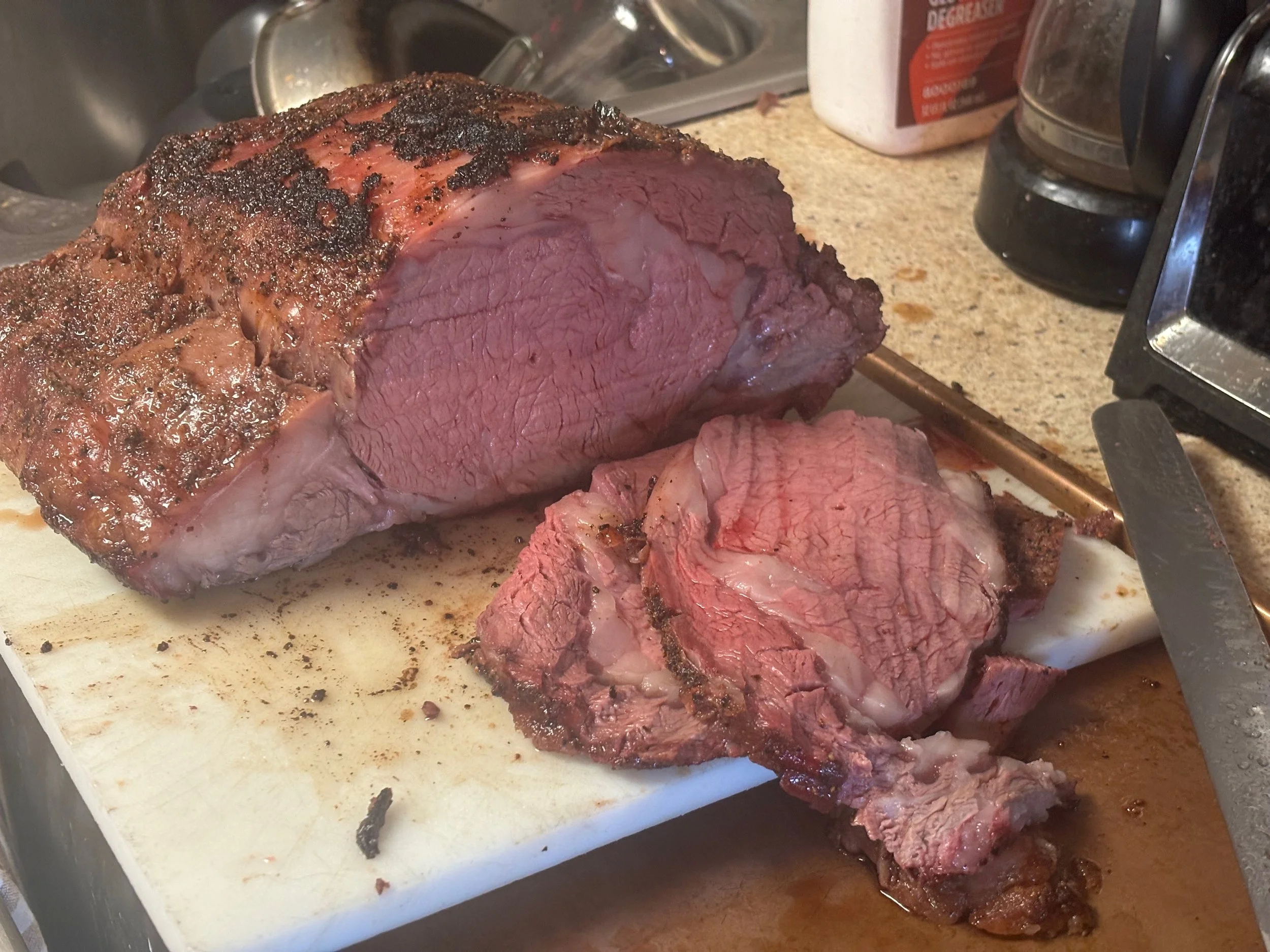 A large cut of cooked beef steak resting on a white cutting board, with some slices cut off and lying next to it, in a kitchen setting.