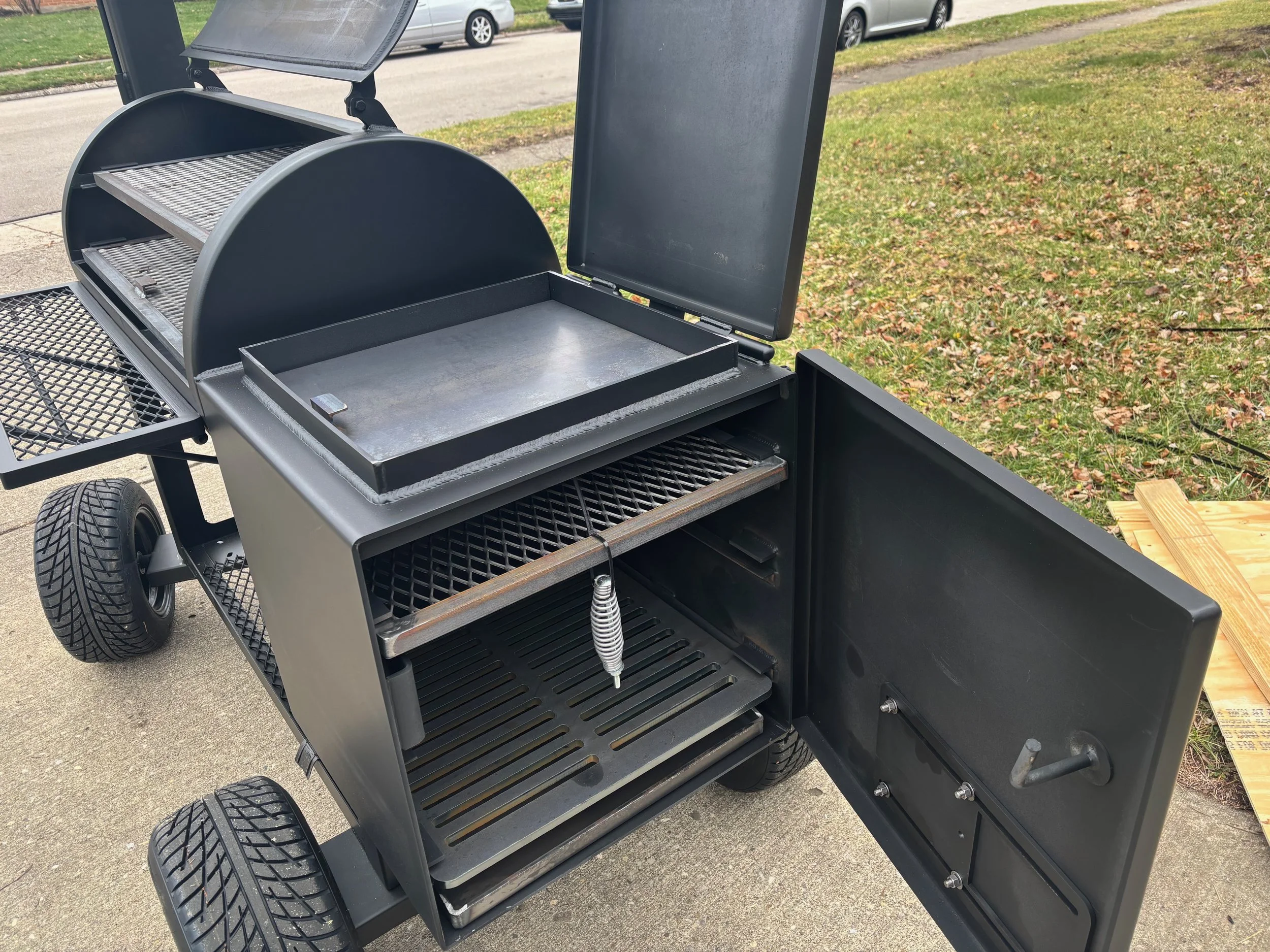 Black outdoor grill with open lid, various racks, and a side tray, situated on a pavement with grass and parked cars in the background.