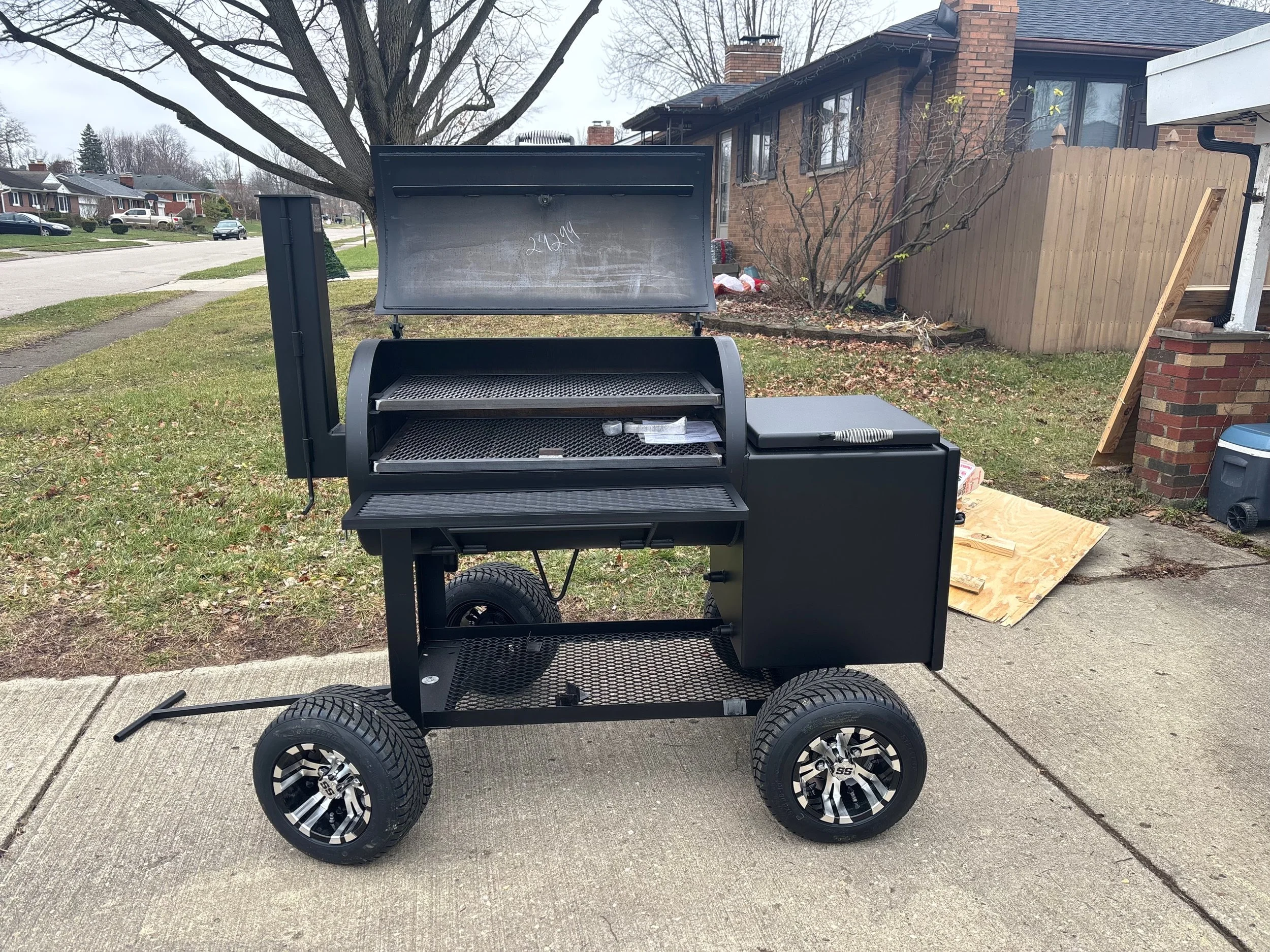 Black portable outdoor grill with wheels, open lid, and attached storage on a concrete sidewalk in a residential neighborhood.
