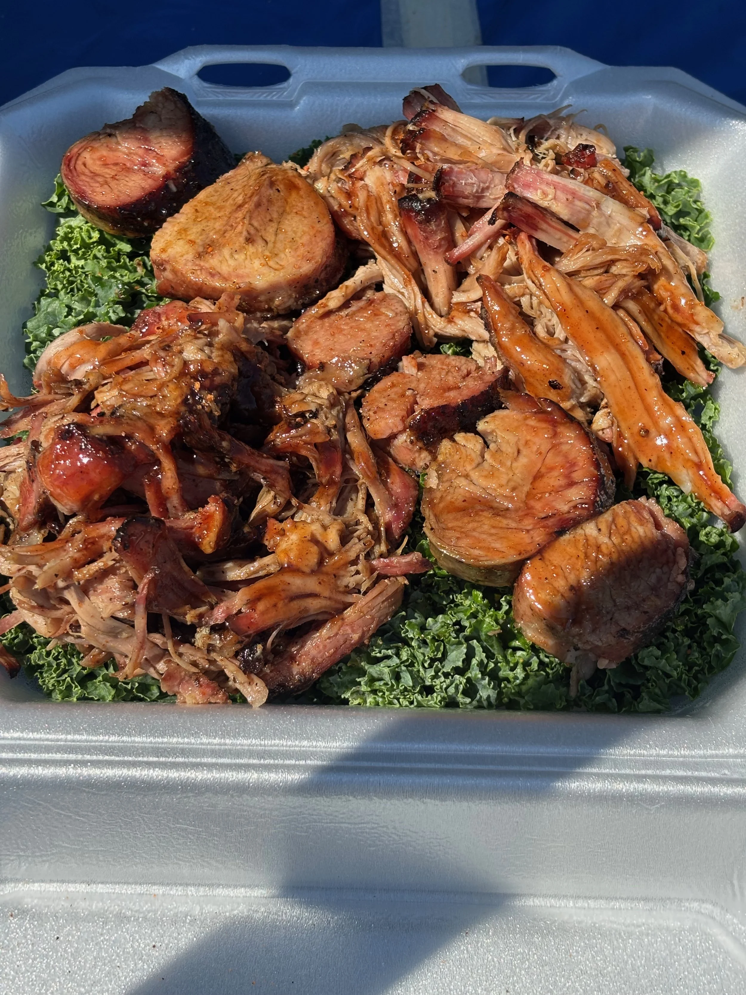 Barbecue tray with sliced brisket, pulled pork, and smoked sausage on a bed of kale.