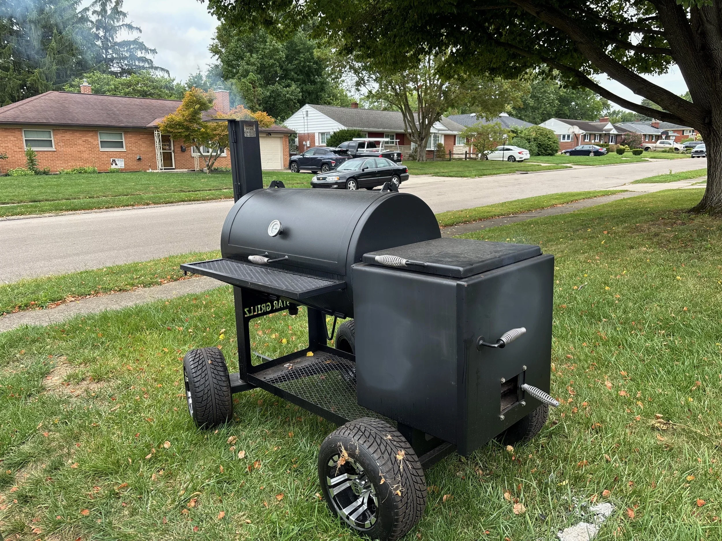 A black portable outdoor smoker grill with wheels, situated on a grassy area next to a sidewalk and tree, with a residential neighborhood in the background.