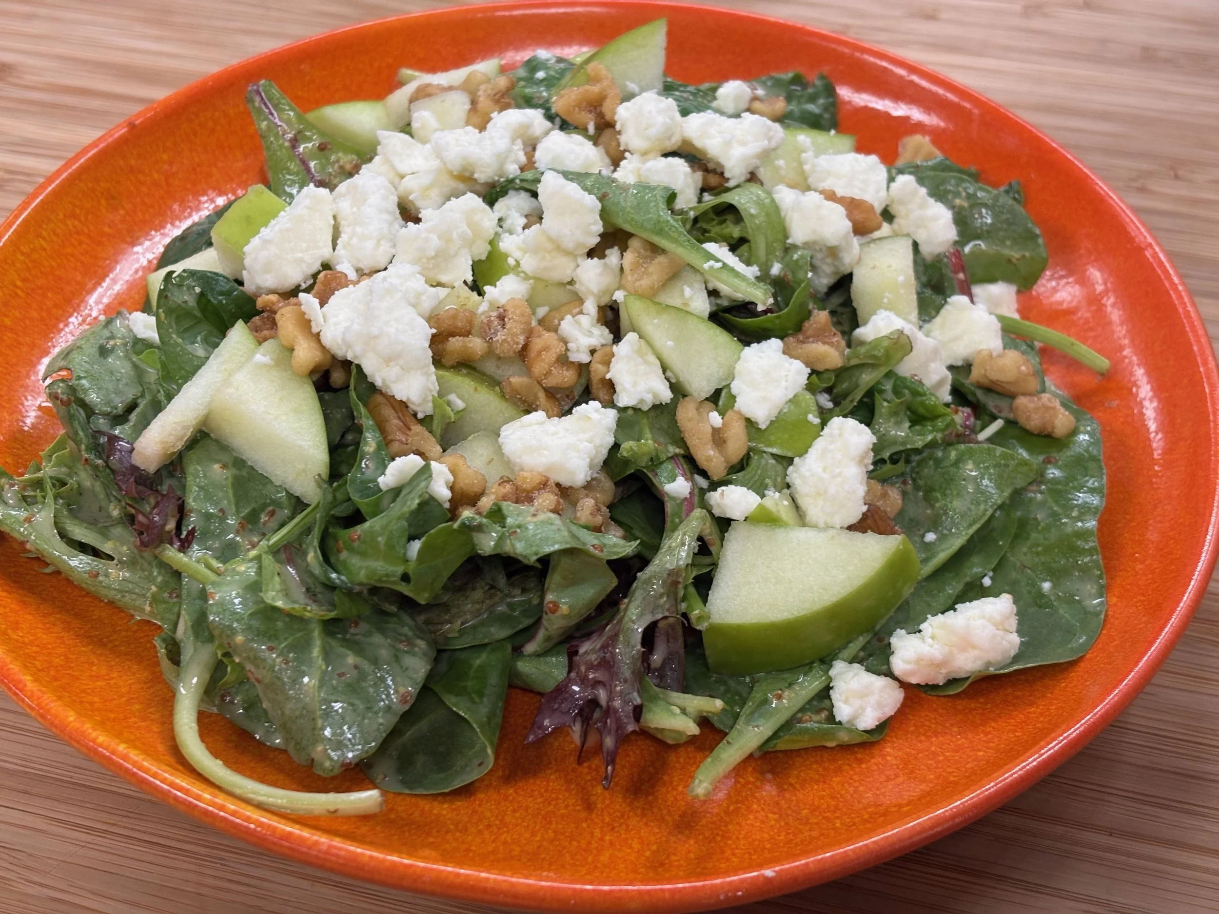 A fresh salad with mixed greens, crumbled white cheese, chopped green apples, walnuts, and sliced cucumber on an orange plate.