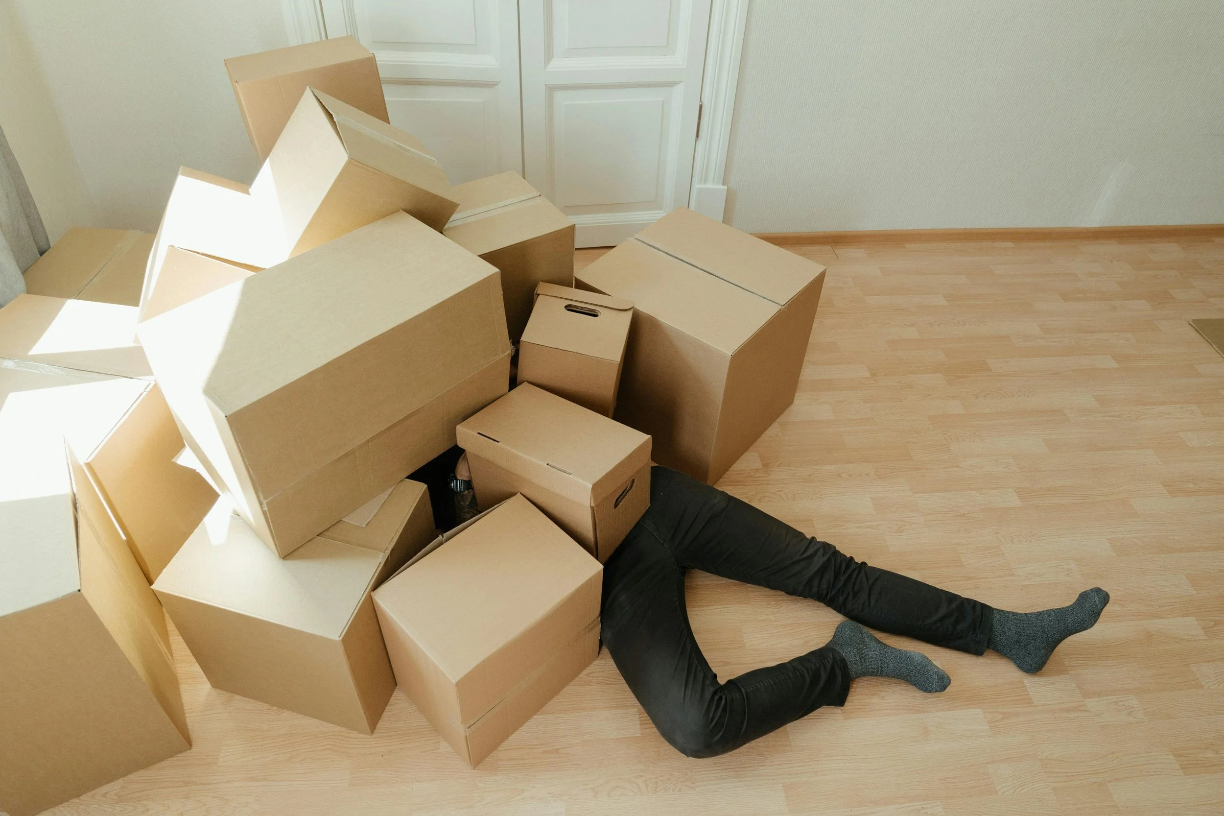 Person lying on the floor with only their legs visible, surrounded by several cardboard boxes in a room with light-colored wood flooring and a closed white door.