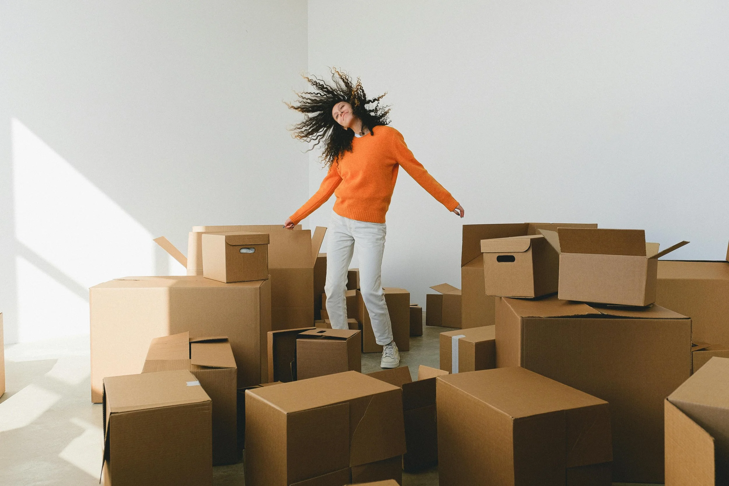 Woman with curly hair dancing or jumping among cardboard boxes in a minimalist room with white walls.