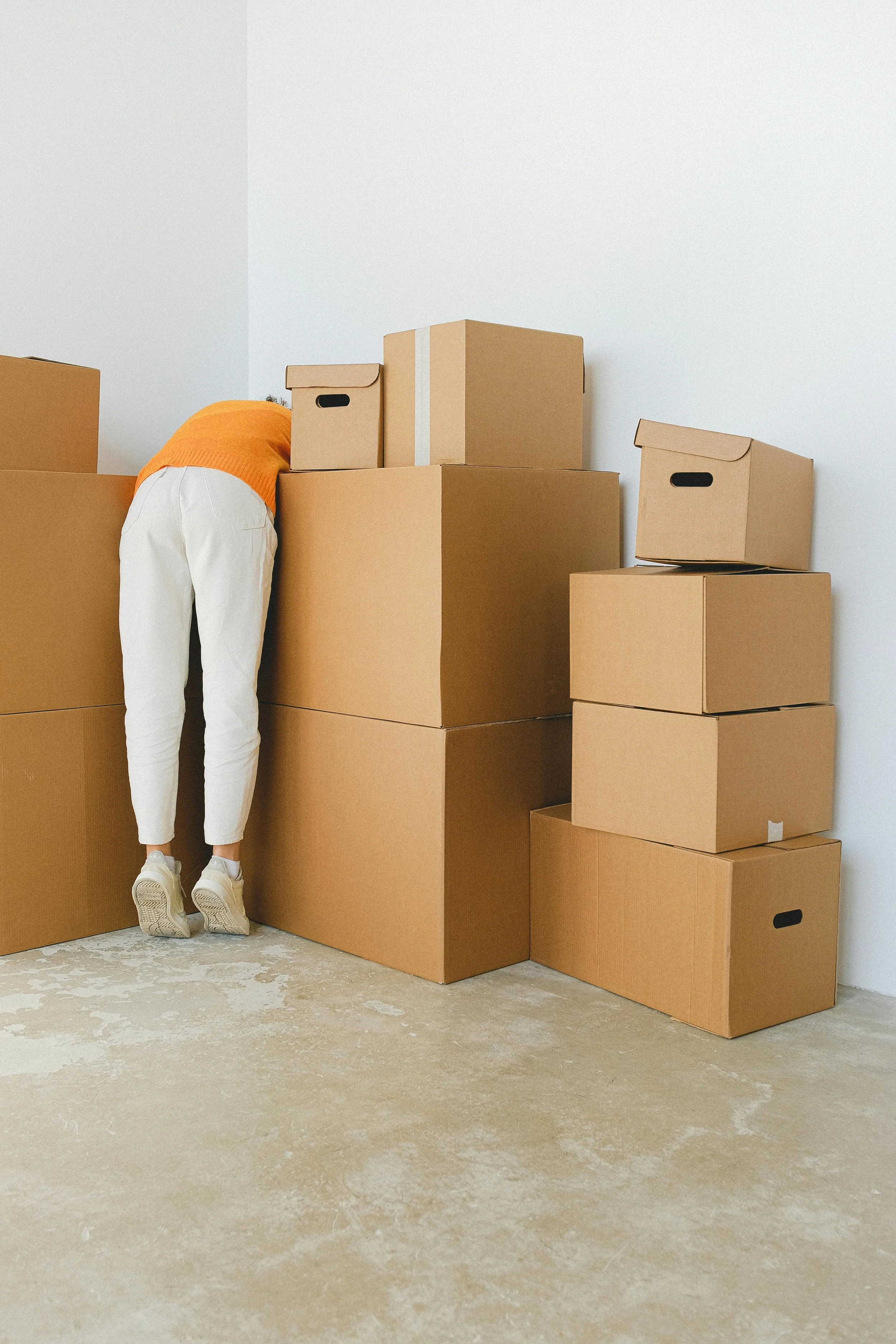 Person wearing orange top and white pants leaning over a pile of cardboard boxes in a room with white walls and a beige floor.