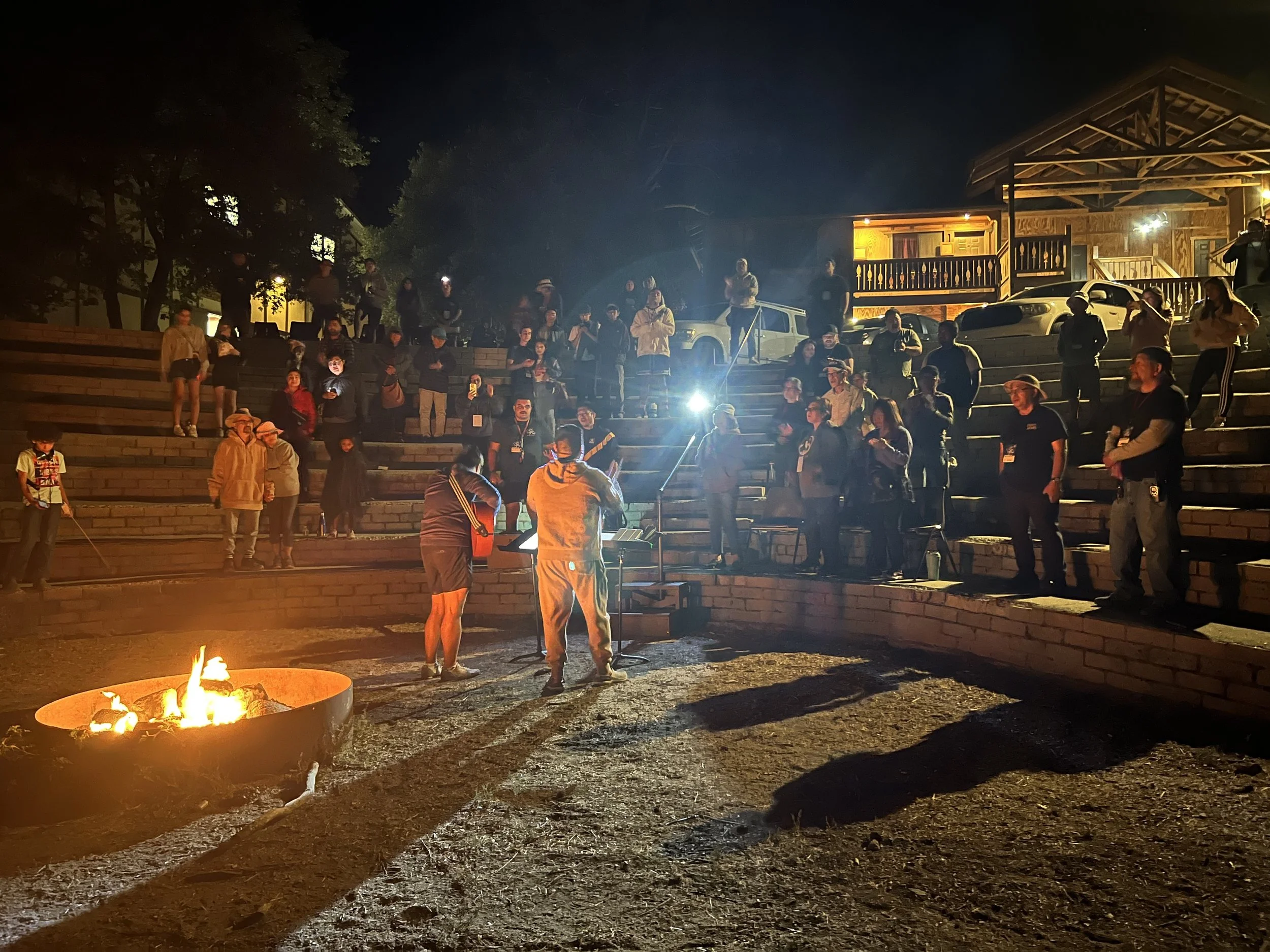 Group of people gathered around a fire pit at night, with some standing or sitting on steps, and a wooden building illuminated in the background.
