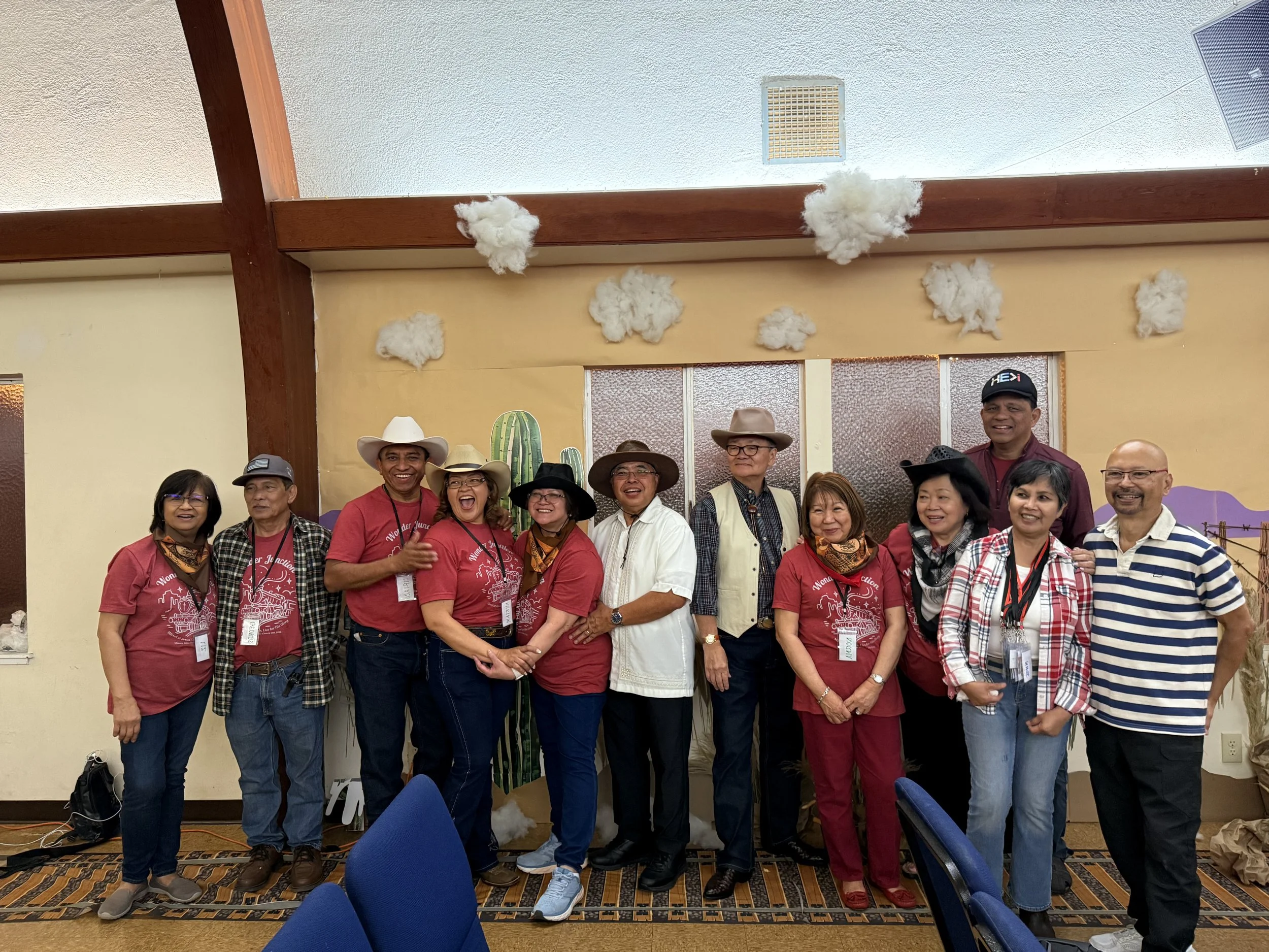 Group of people standing together indoors, some wearing wide-brimmed hats and red T-shirts, smiling and posing for a photo in front of a wall with decorative elements and tissue paper decorations.