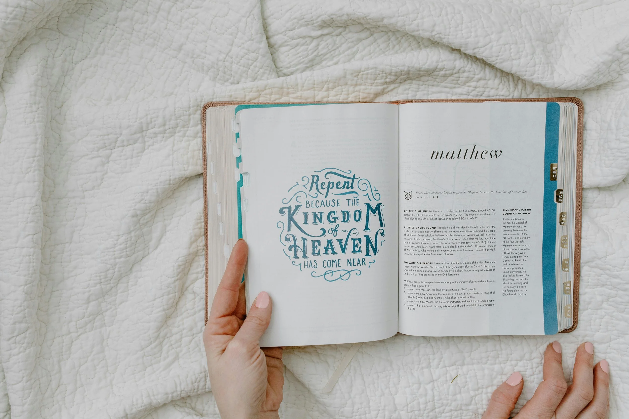Open Bible with pages displaying text, held by a person's hands, resting on a white quilted fabric.