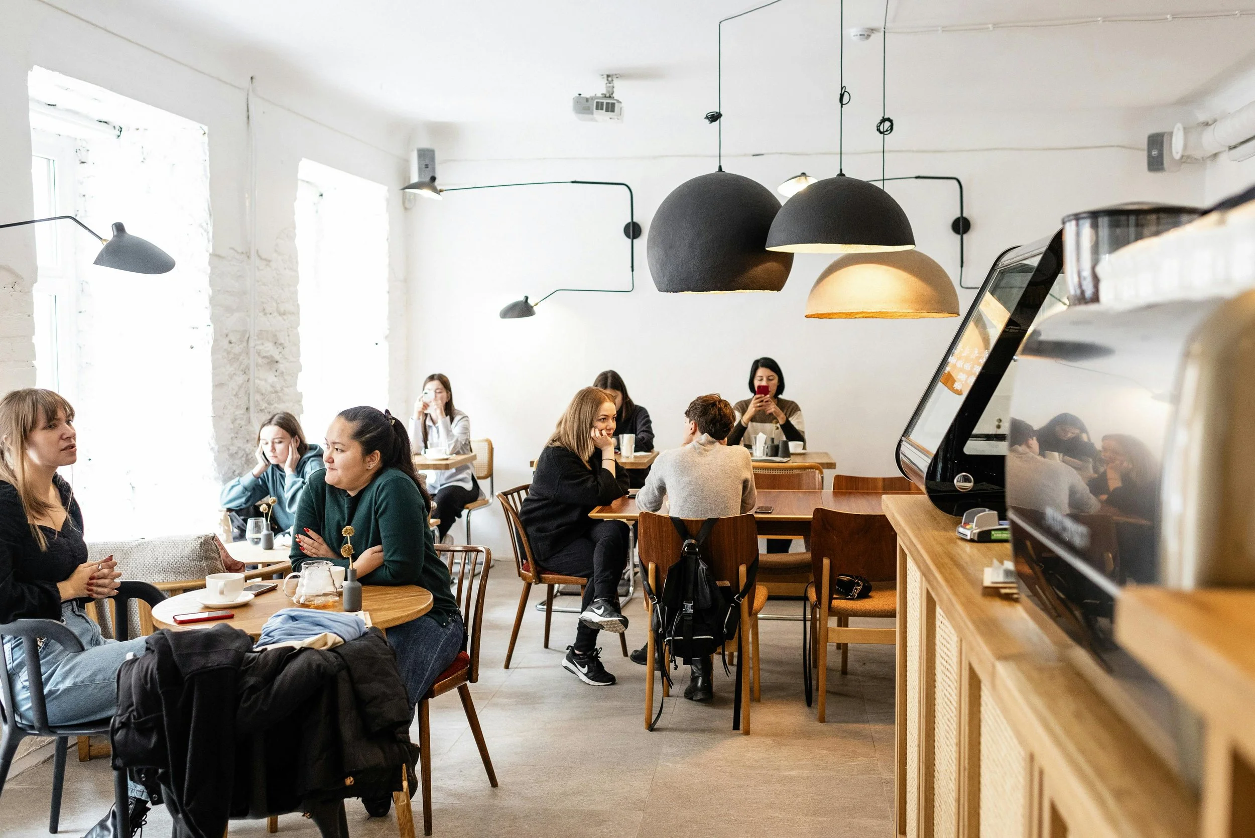 People sitting at tables in a cozy coffee shop with white brick walls, large windows, and modern pendant lights hanging from the ceiling.