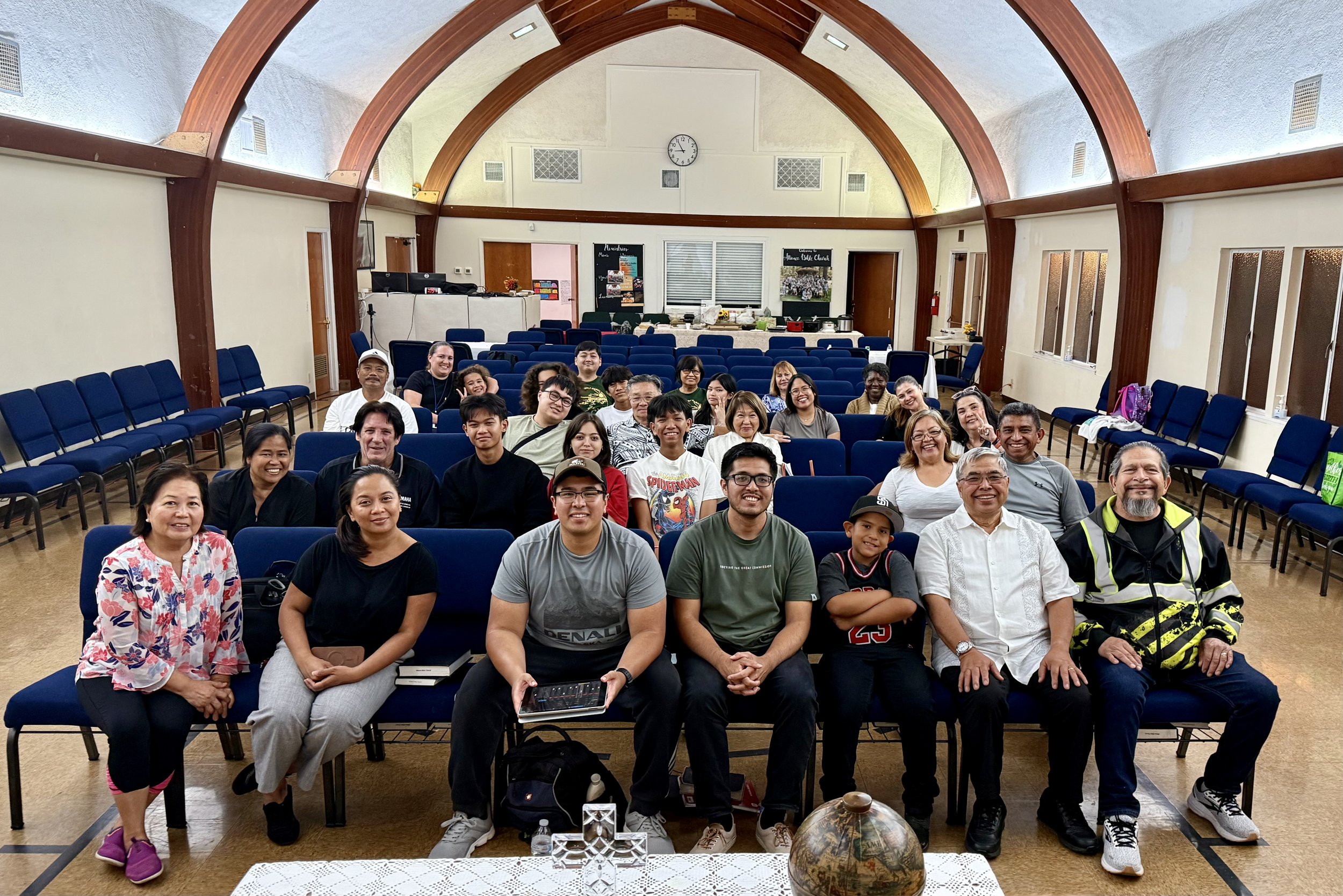 A group of diverse people posing for a photo inside a church with wooden arches, seating for attendees, and a clock on the wall.