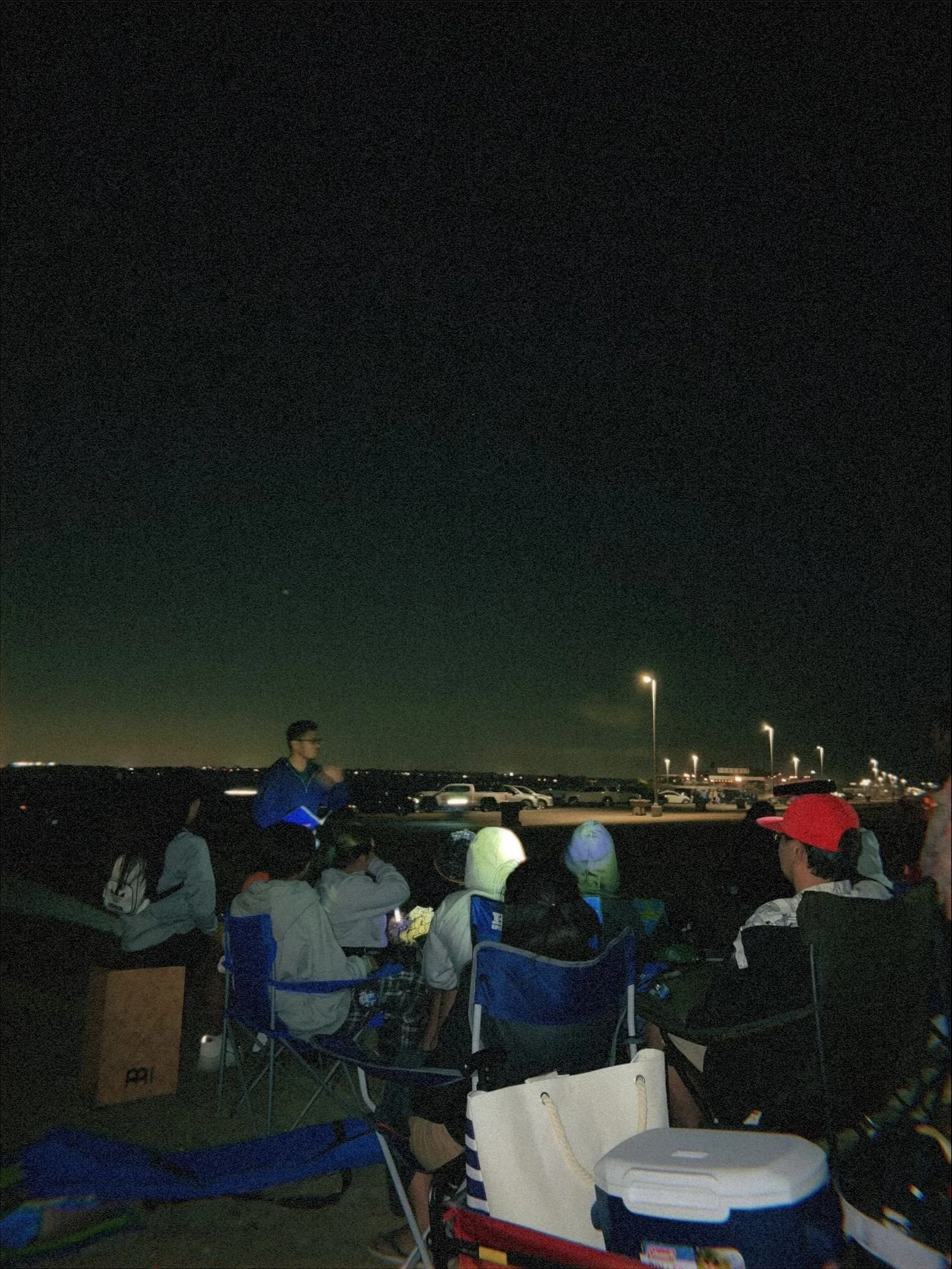 People sitting outdoors at night, watching fireworks in the sky. Some are taking photos or videos. There are parked cars and streetlights in the background.