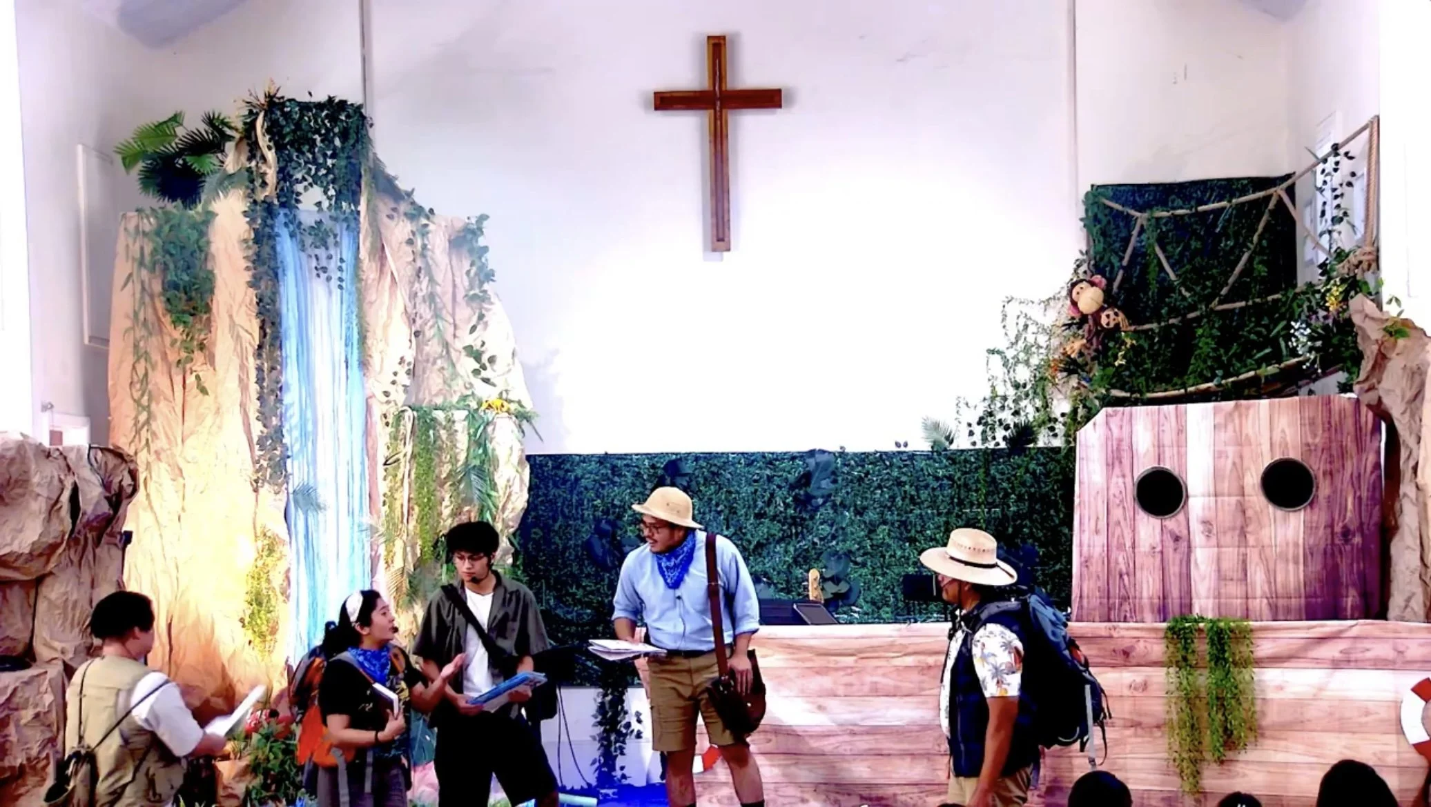 People gathered in front of a stage decorated with greenery, wood, and crosses, attended by children and adults, in a church or community setting.