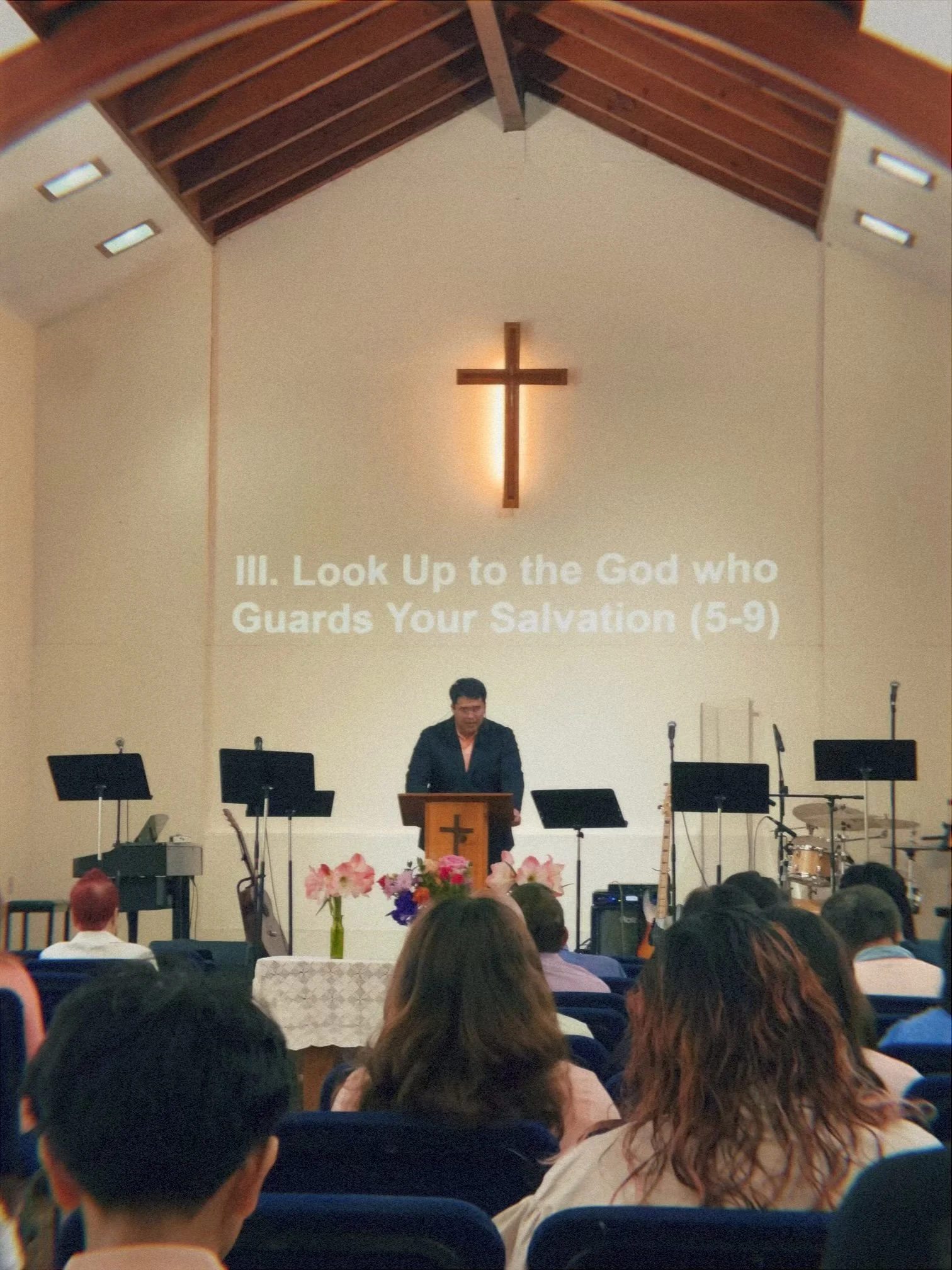 A church service with a man standing at a pulpit, a large wooden cross illuminated on the wall behind him, and an audience seated facing the stage. There are music stands, flowers, and musical instruments on the stage.
