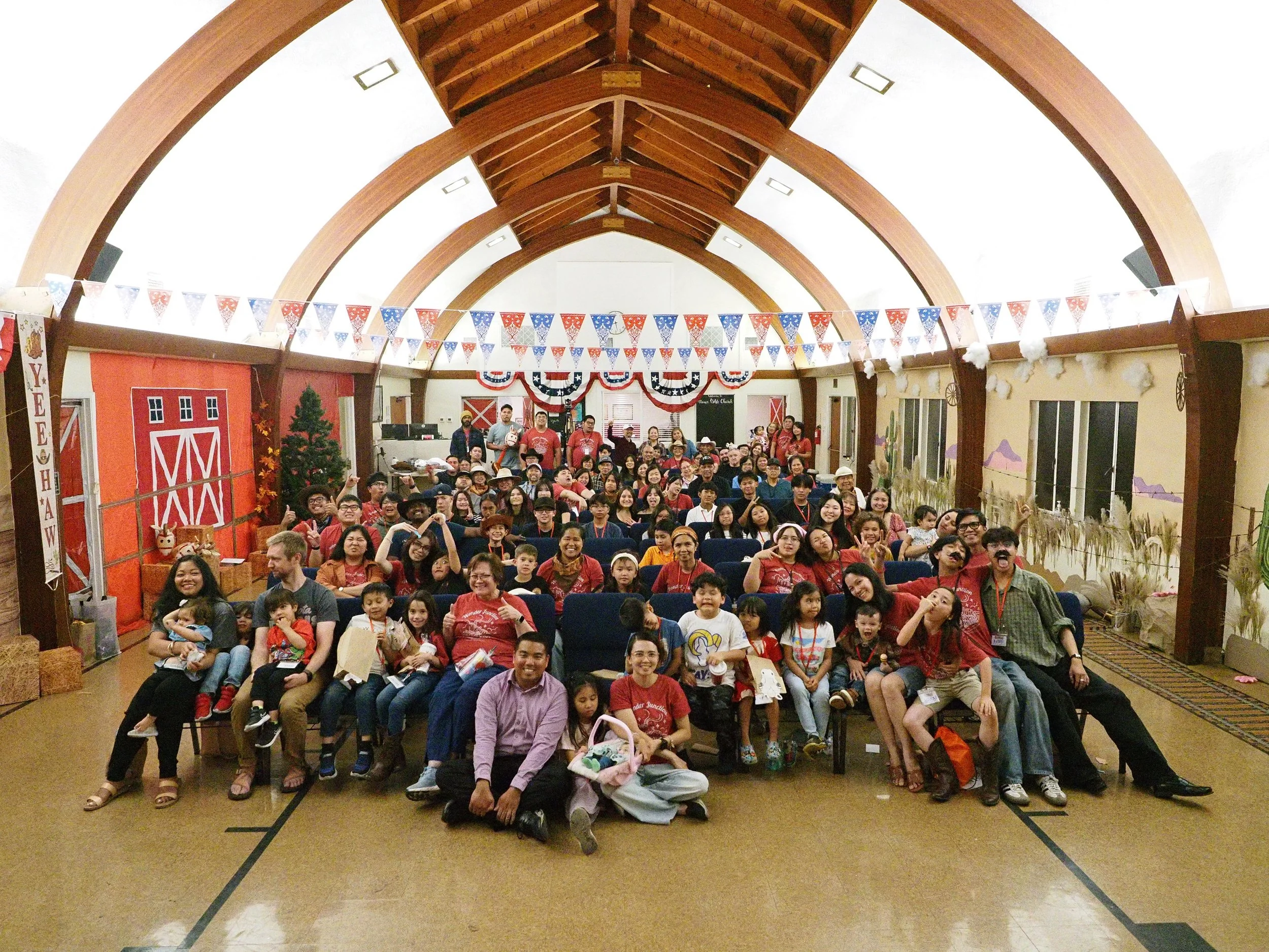 A group of people gathered inside a hall decorated with red, white, and blue banners and bunting, with a wooden arch ceiling. Many attendees are smiling and sitting in rows of chairs, some with children. There are decorative elements like a fake barn, a small Christmas tree, and wall murals with a rural theme.