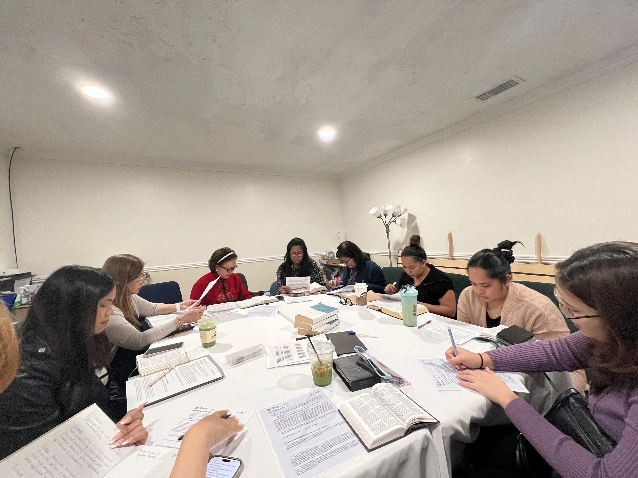 Group of women sitting around a long table engaged in reading and discussion, with open books, papers, and drinks on the table in a room with plain walls and ceiling lights.