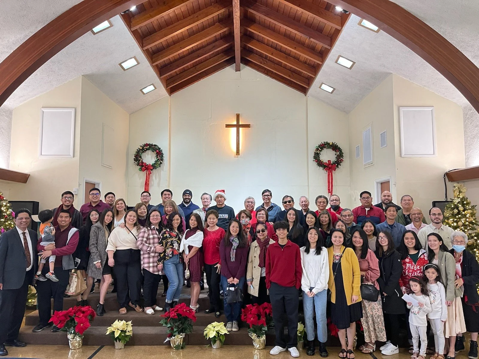 A large group of people gathered in a church decorated for Christmas, with wreaths, poinsettias, and a Christmas tree, under a wooden ceiling with a cross on the wall.