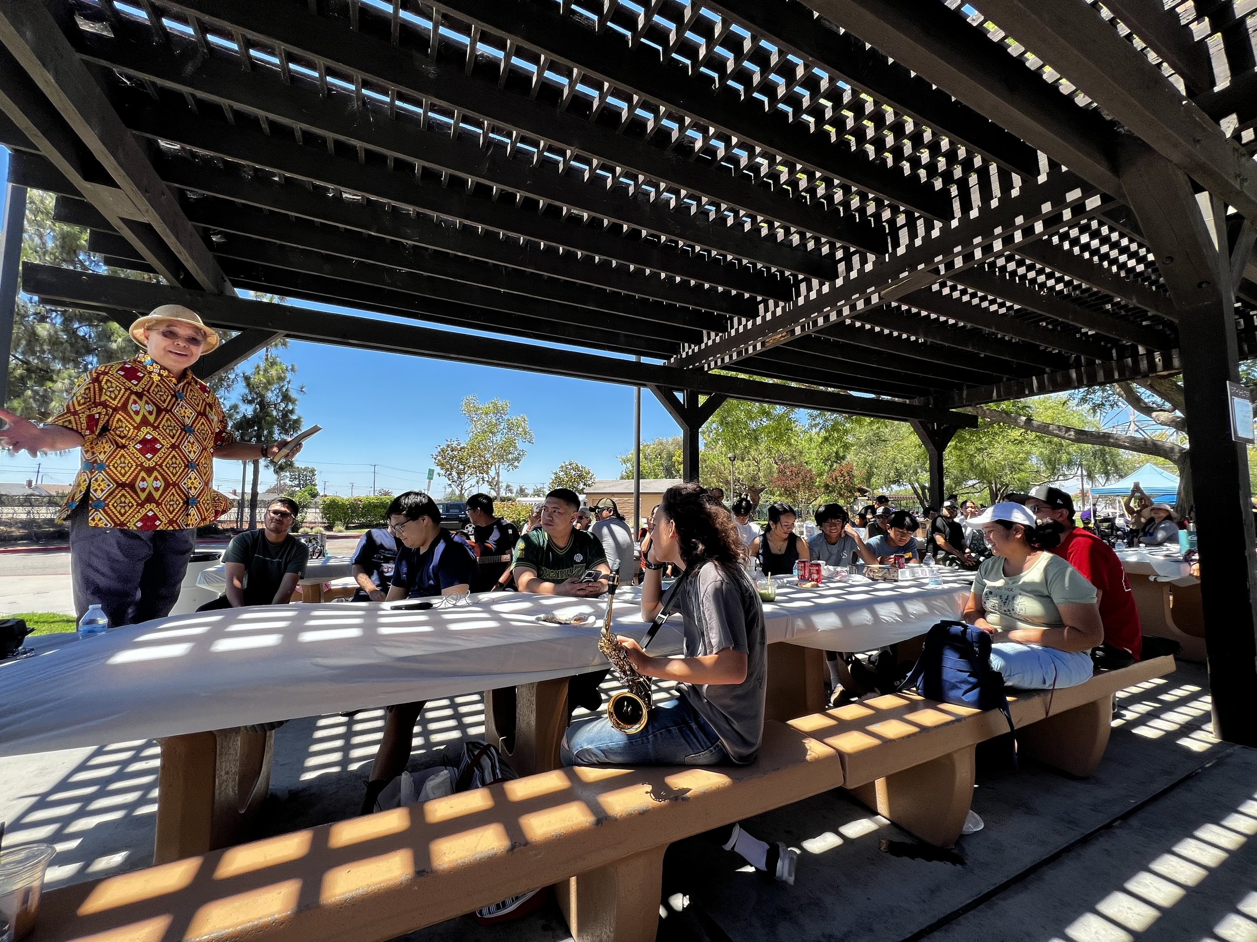 A group of people sitting at picnic tables under a large wooden pavilion outdoors on a sunny day. A woman wearing a colorful patterned shirt, hat, and sunglasses stands and smiles, holding a smartphone. The background includes trees, a blue sky, and some tents in the distance.