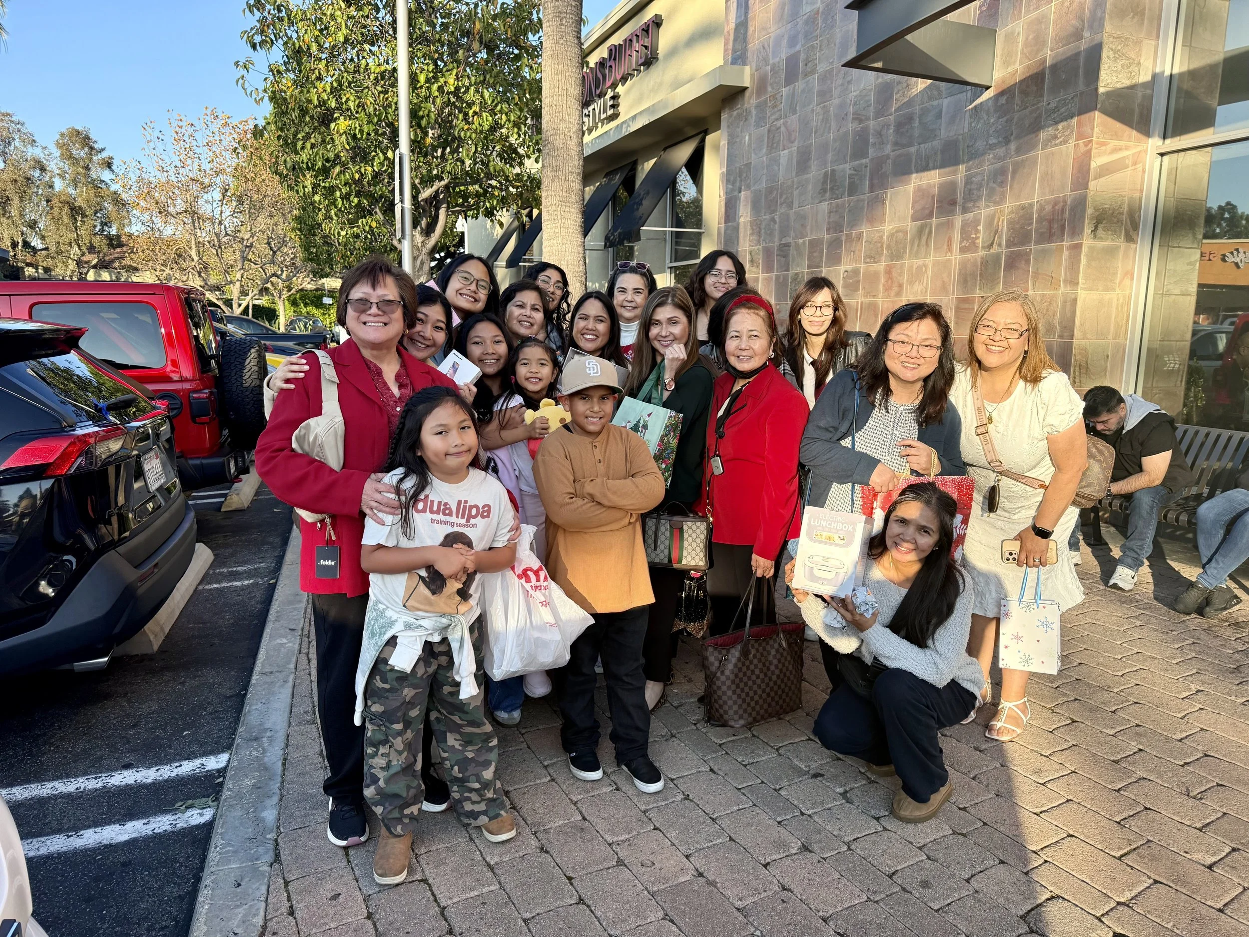 A diverse group of women and children smiling and posing for a photo outside a building with a sign that reads "ONS BUFFET STYLE." They are standing on a sidewalk next to a parking lot with several cars, including a red vehicle. The background features trees and a clear sky.