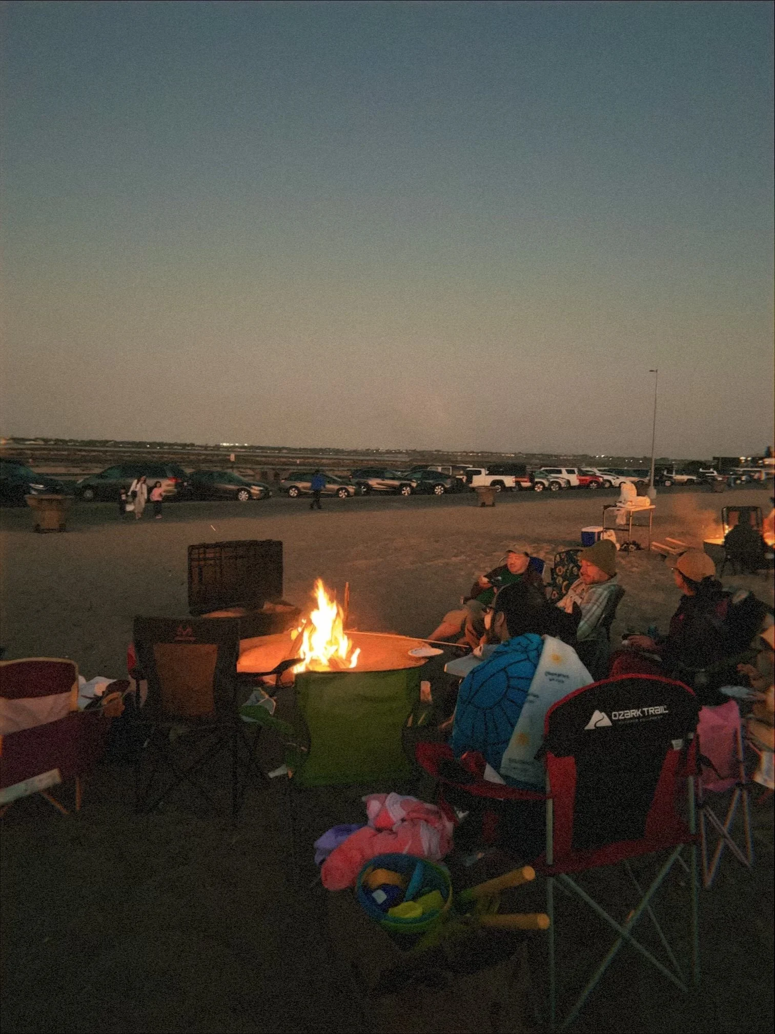 People gathered around a campfire with camping chairs and gear, outdoors during the evening or early night, with a wide open sky and parked cars in the background.