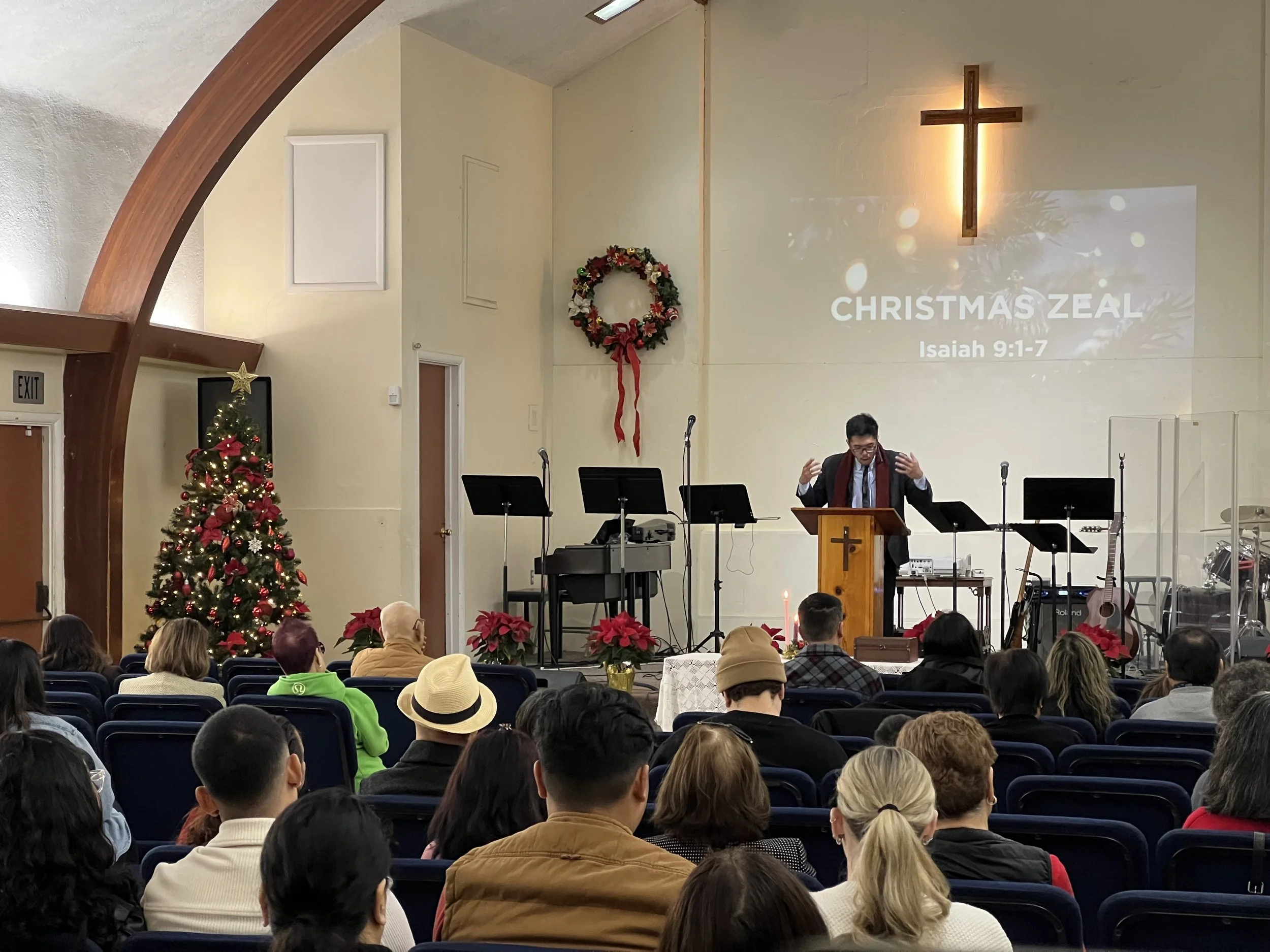 A church service or gathering with many attendees seated and facing the front. The front wall displays a large illuminated cross, a Christmas wreath, and a projected slide that reads "Christmas Zeal Isaiah 9:1-7." There is a Christmas tree decorated with red ornaments and lights to the left, along with poinsettias placed at the front. A man is speaking at a wooden pulpit.