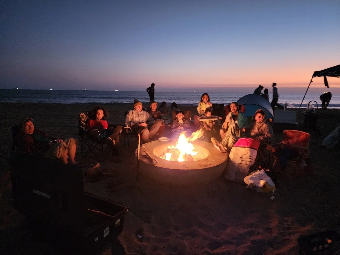 People gathered around a campfire on a beach at sunset, sitting on chairs, with tents and the ocean in the background.