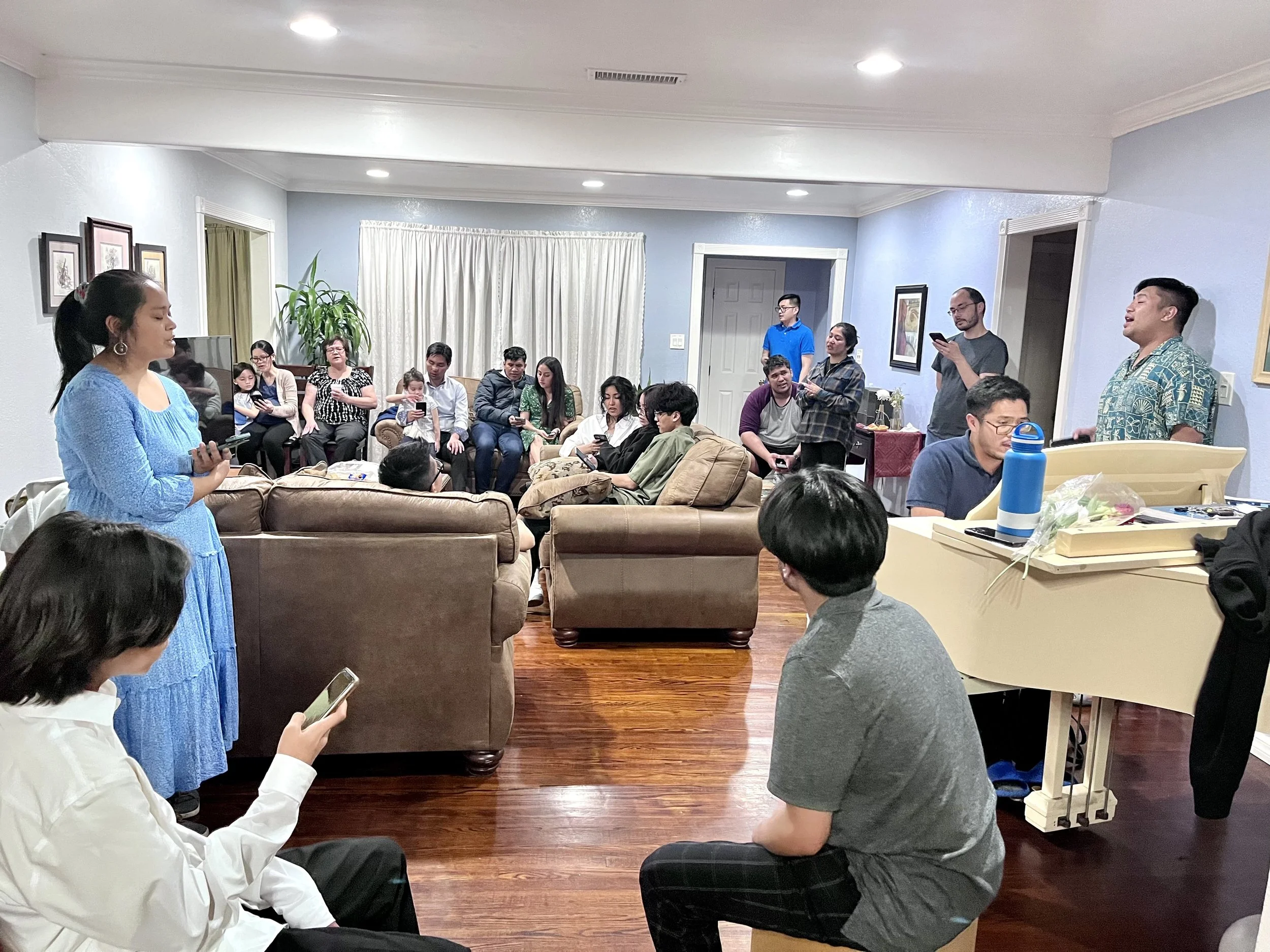 A group of people gathered in a living room, some standing and others sitting on sofas, engaging in a social or family gathering.