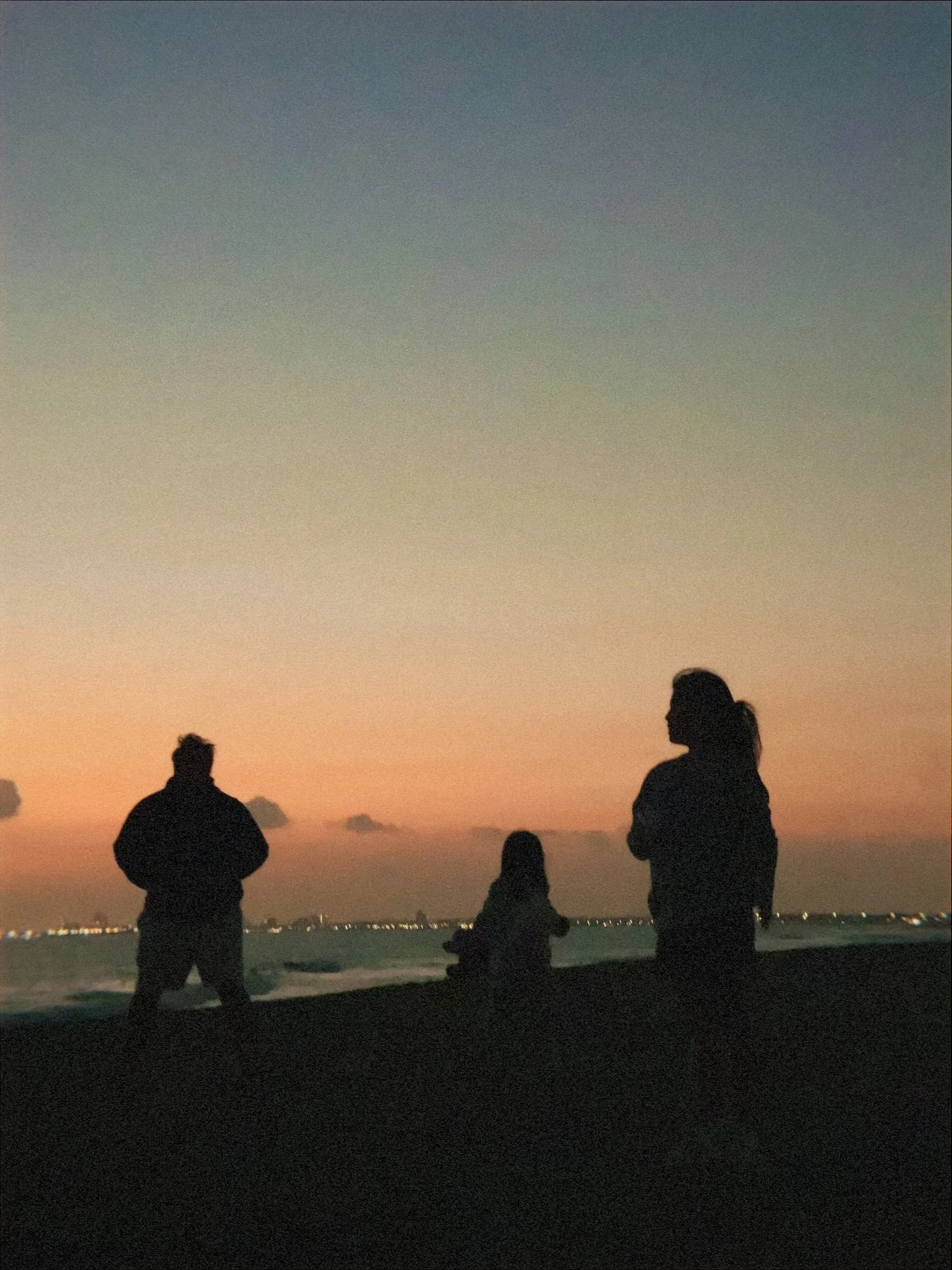 People standing on a beach at sunset, with a calm sea and city lights in the distance.