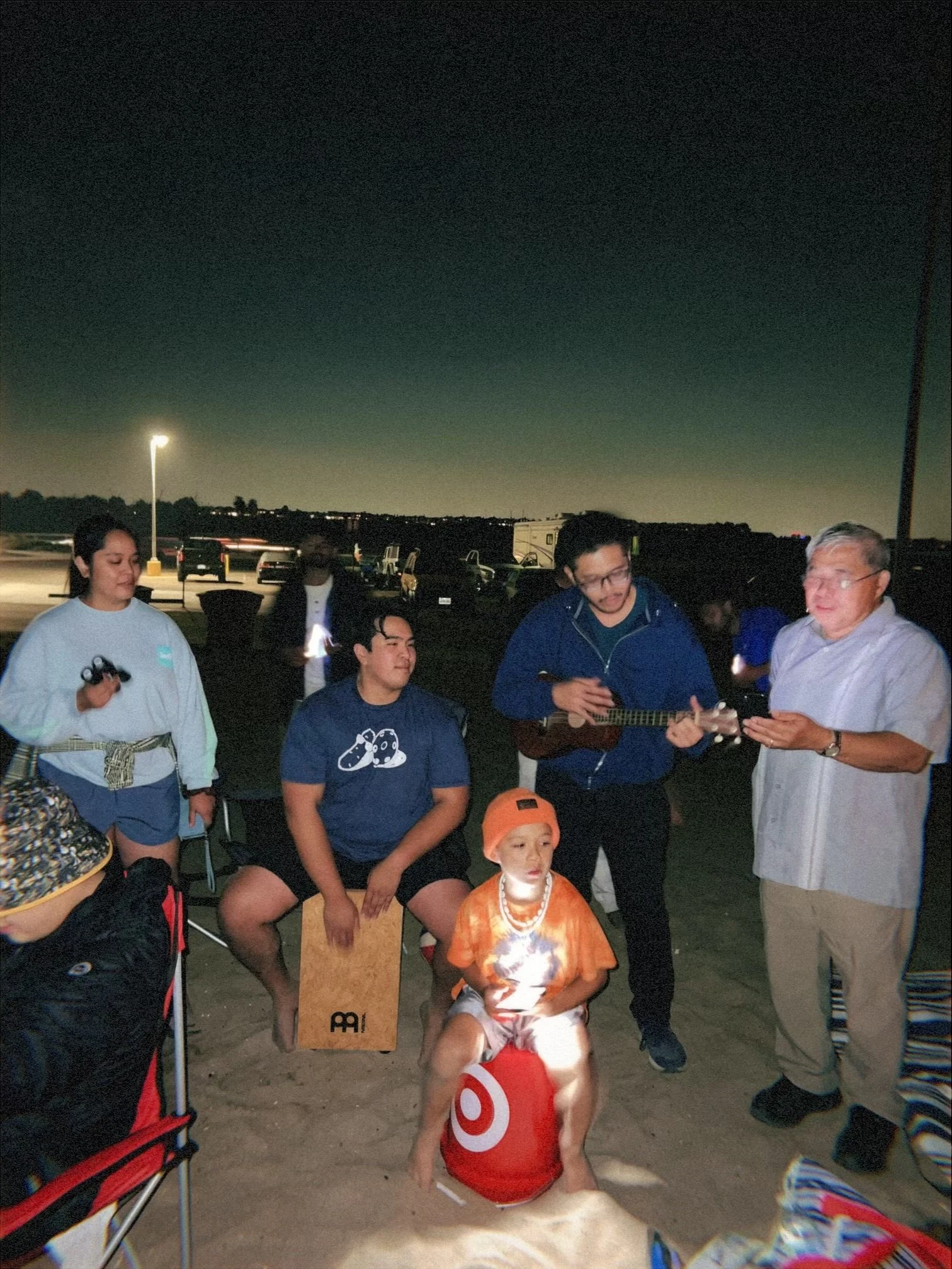 A group of people gathered outdoors at night, with a dark sky and few streetlights in the background. Some people are standing and others are sitting, including a young child in a red Target shirt and a woman holding a smartphone.
