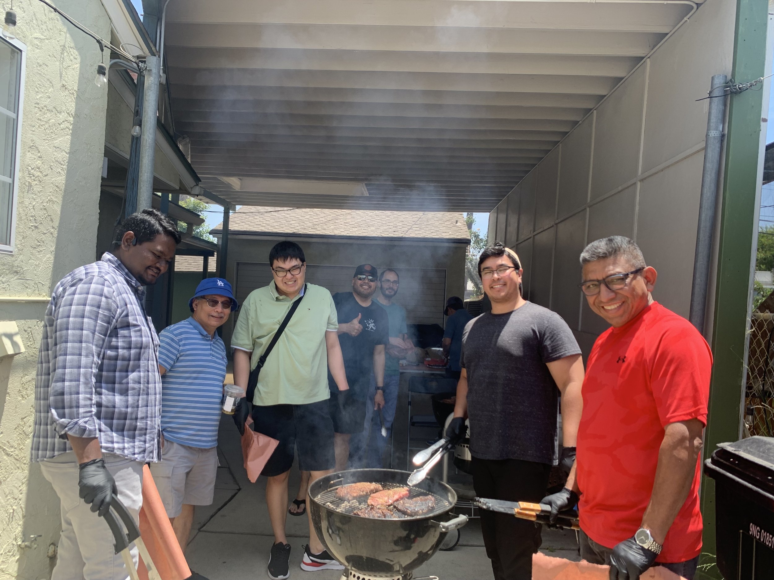 A group of men cooking on a barbecue grill under a patio cover in a backyard, smiling and enjoying themselves.