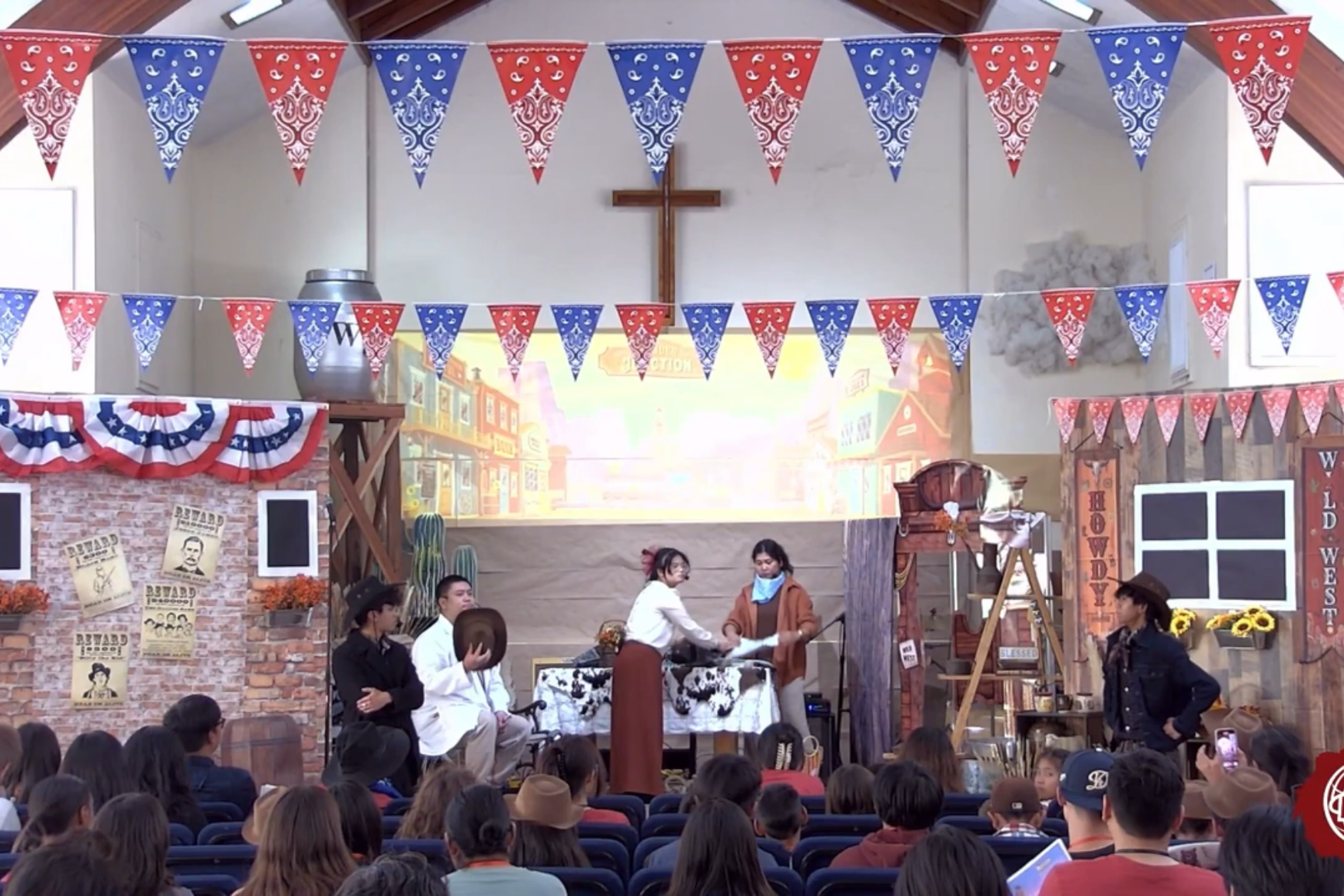 Stage set with Western and patriotic decorations, including red, white, and blue bunting and banners. Actors in cowboy costumes perform in front of a backdrop depicting a Western town. Audience members watch attentively.