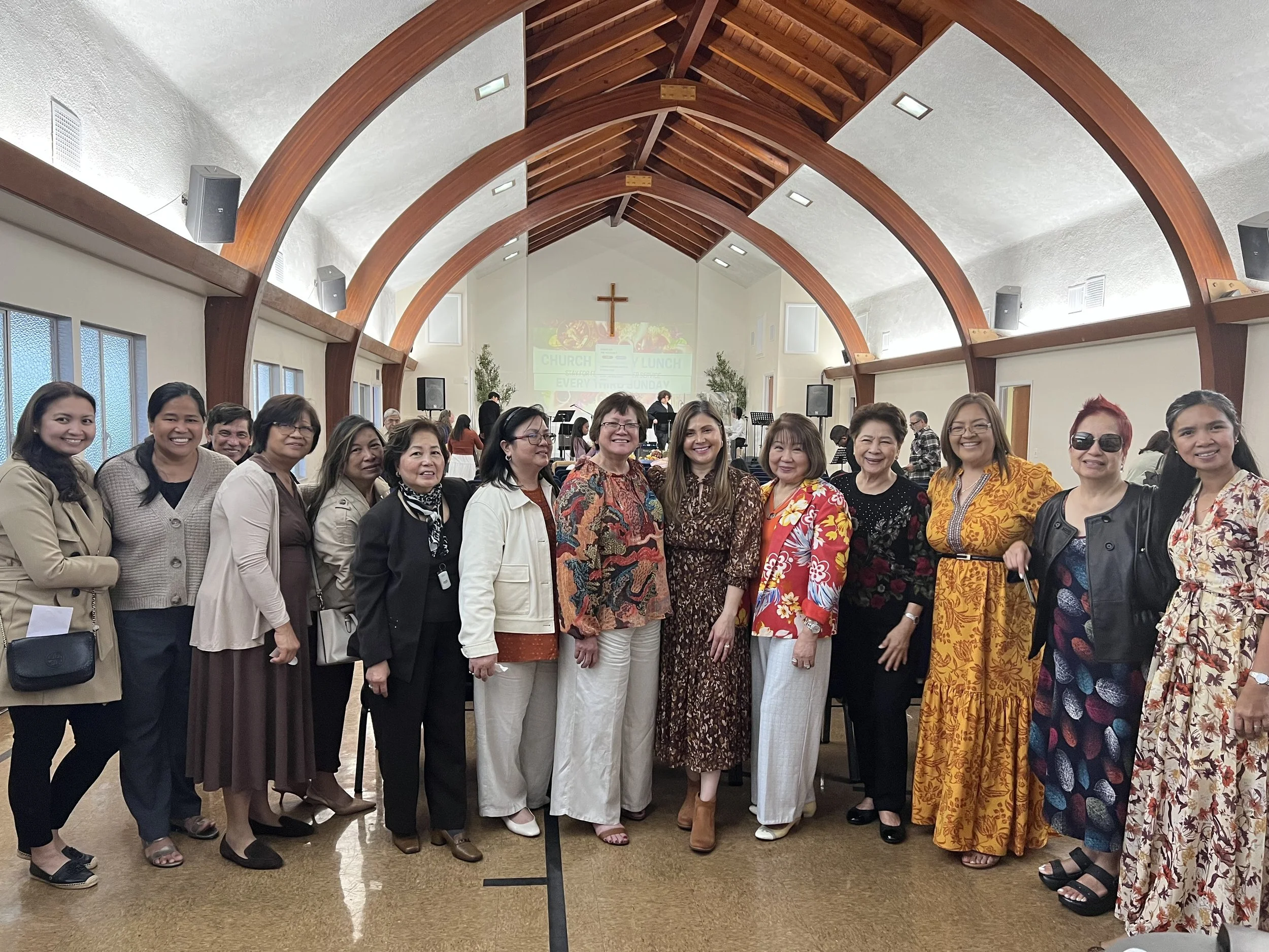 Group of women standing inside a church with wooden arch ceilings, a cross on the wall, and a projection screen displaying church info.