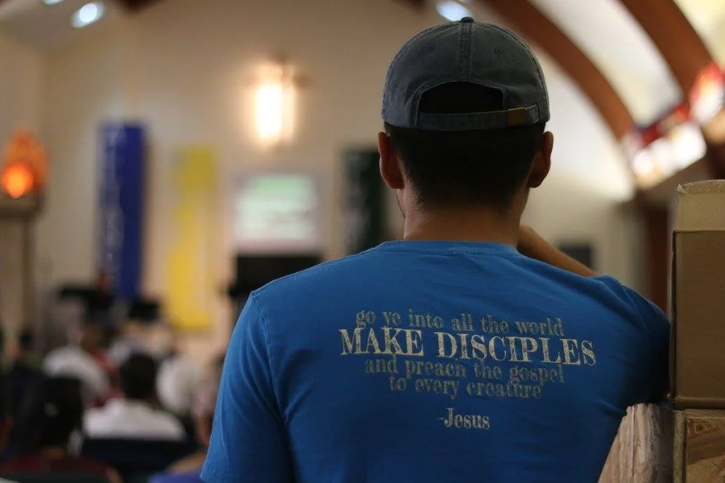 A person wearing a blue T-shirt with the text "Go into all the world, MAKE DISCIPLES and preach the gospel to every creature - Jesus" on the back, inside a church or auditorium, with other people and a projector screen in the background.
