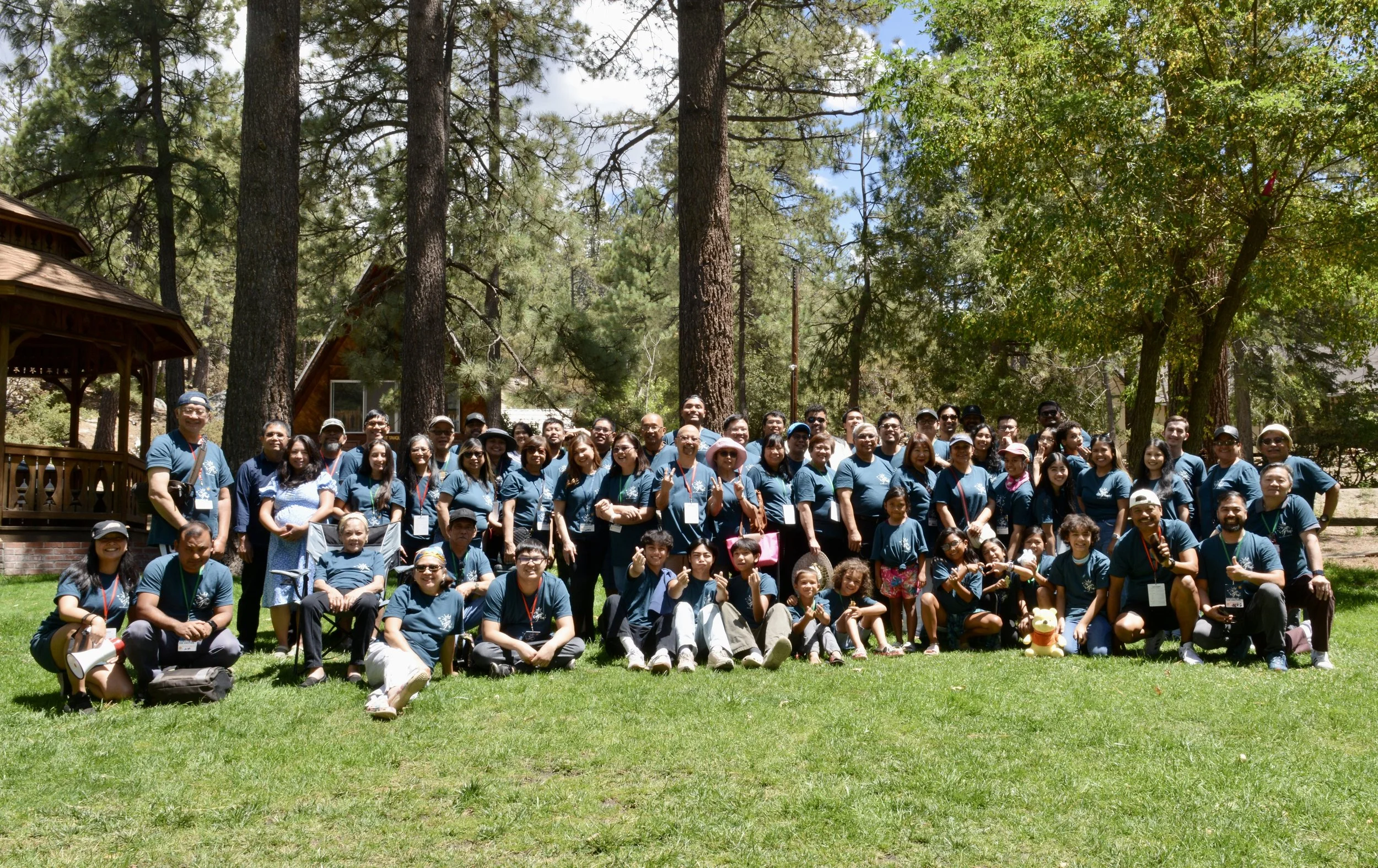 Group of people outdoors in a forested park, posing for a photo, with trees and blue sky in the background.
