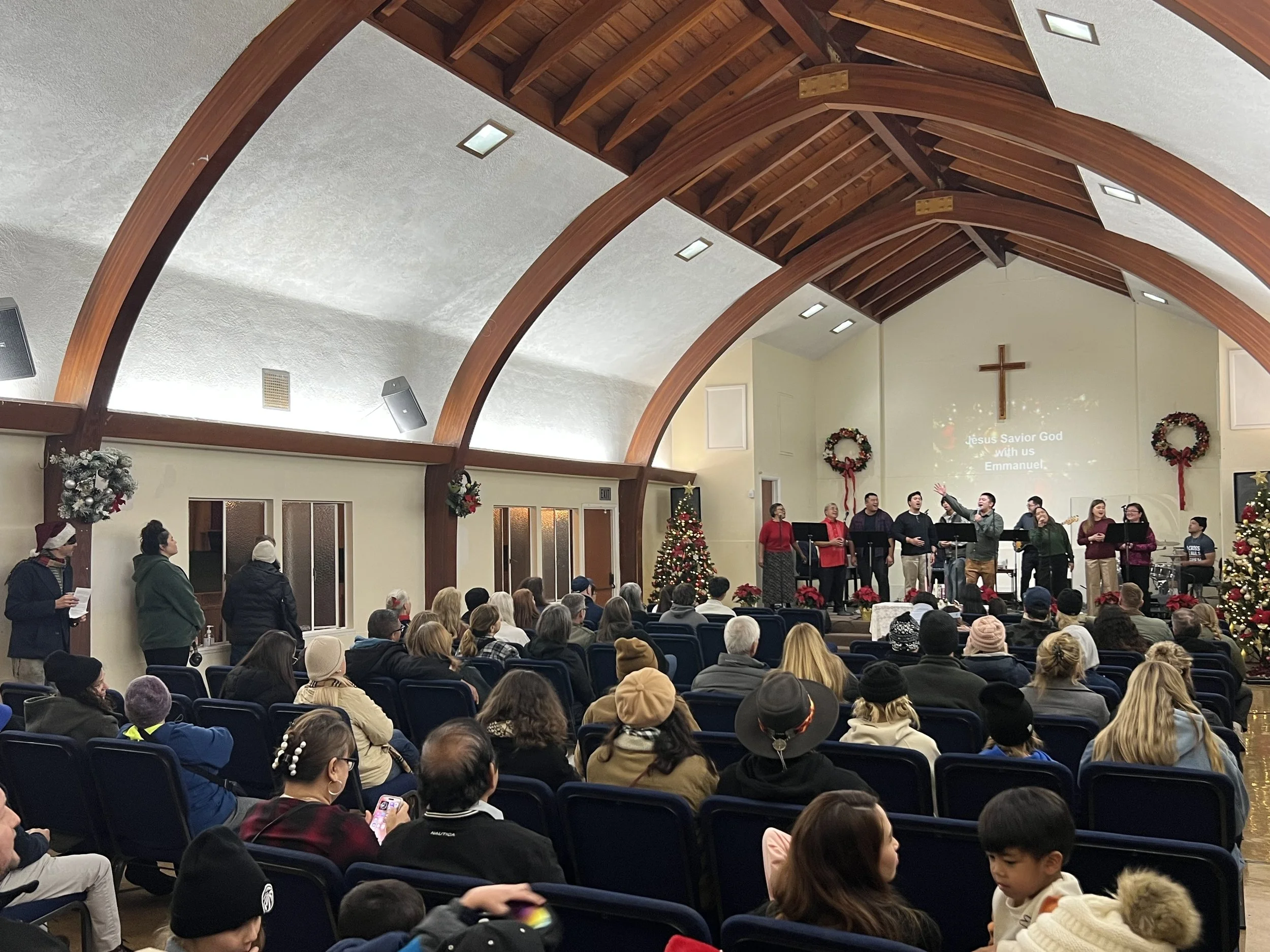 Inside of a church decorated for Christmas, with a choir singing on stage, Christmas wreaths, and a projected message about Jesus Christ from the choir room. Audience members seated, some wearing winter clothing.
